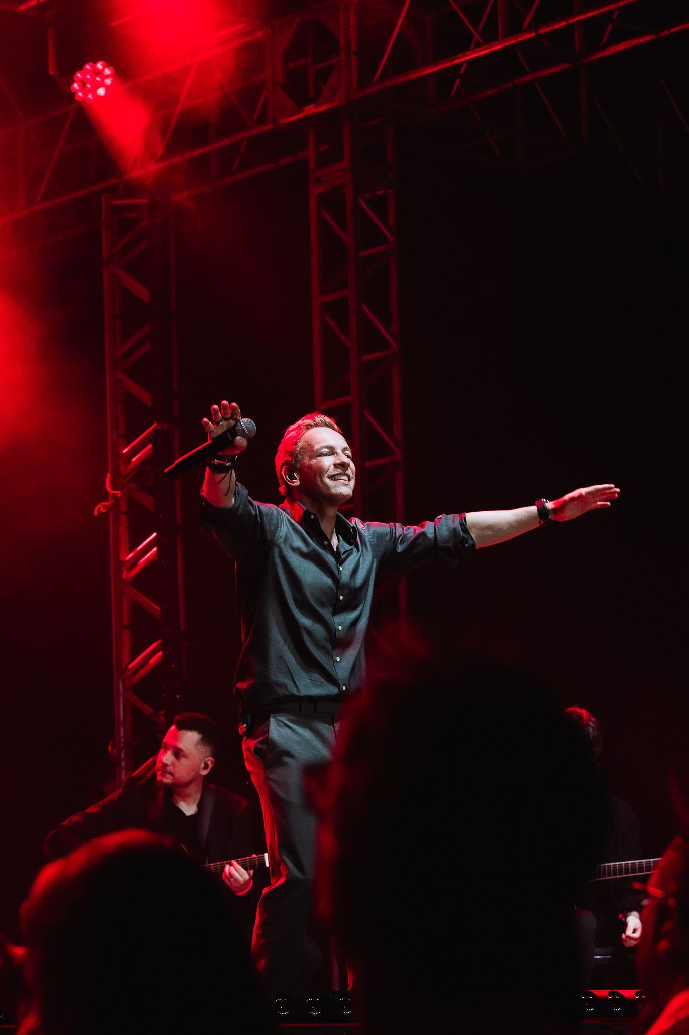 Cantor se apresentando em um palco iluminado em vermelho com os braços estendidos, banda visível ao fundo.