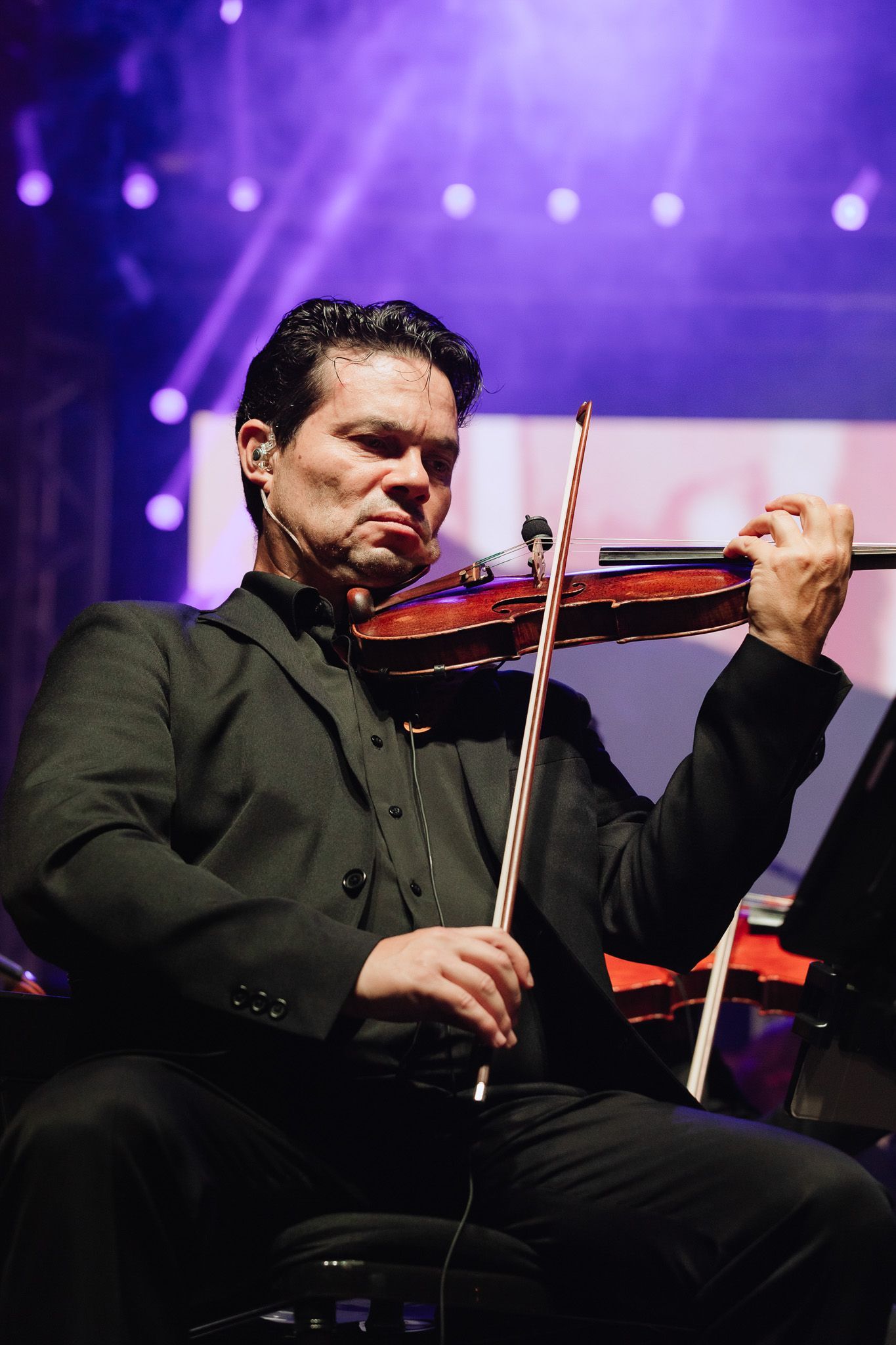 Violinista se apresentando no palco sob luzes roxas, tocando intensamente em um traje preto.