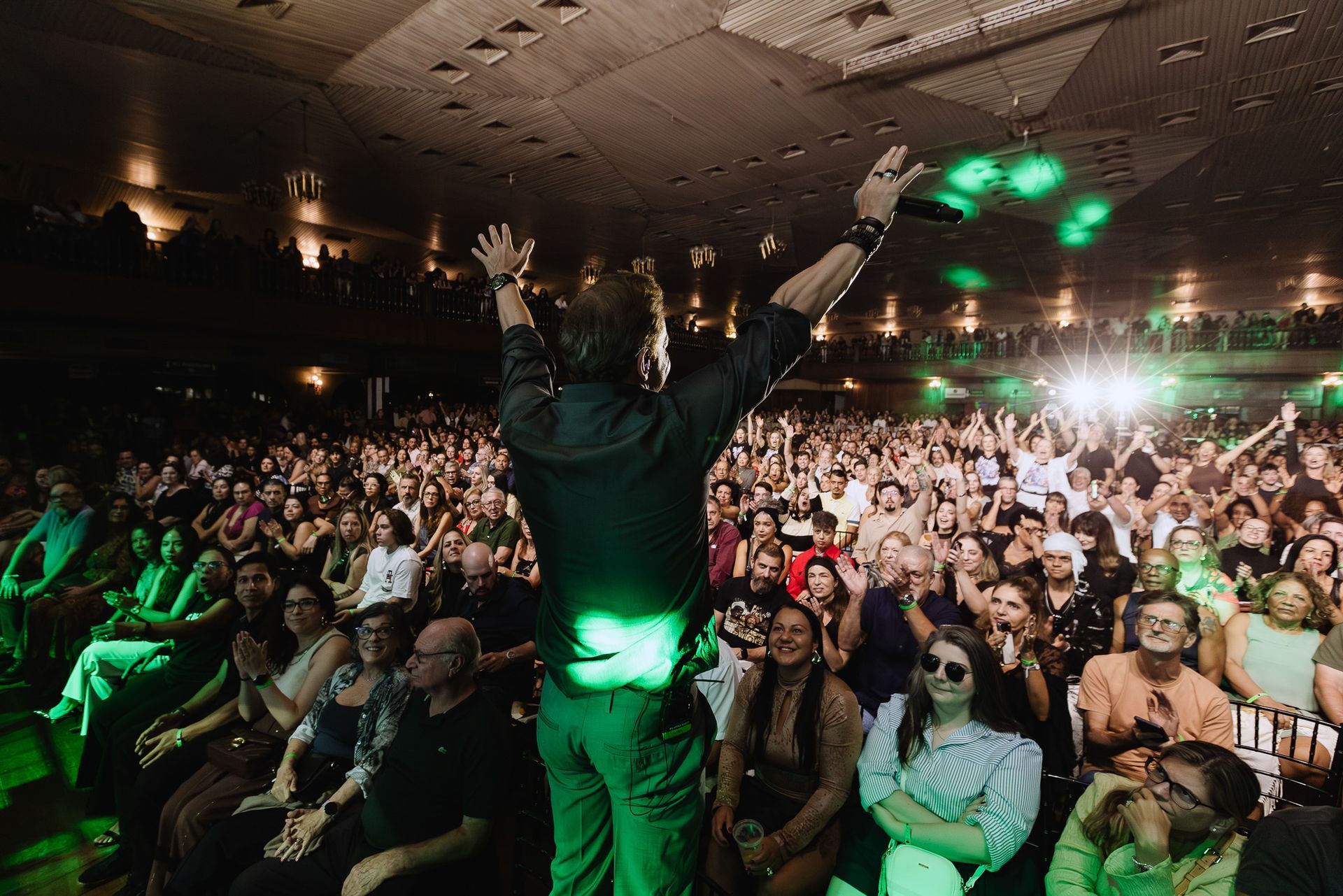 Artista de braços erguidos diante de uma plateia lotada em uma sala de concertos com iluminação verde tênue.