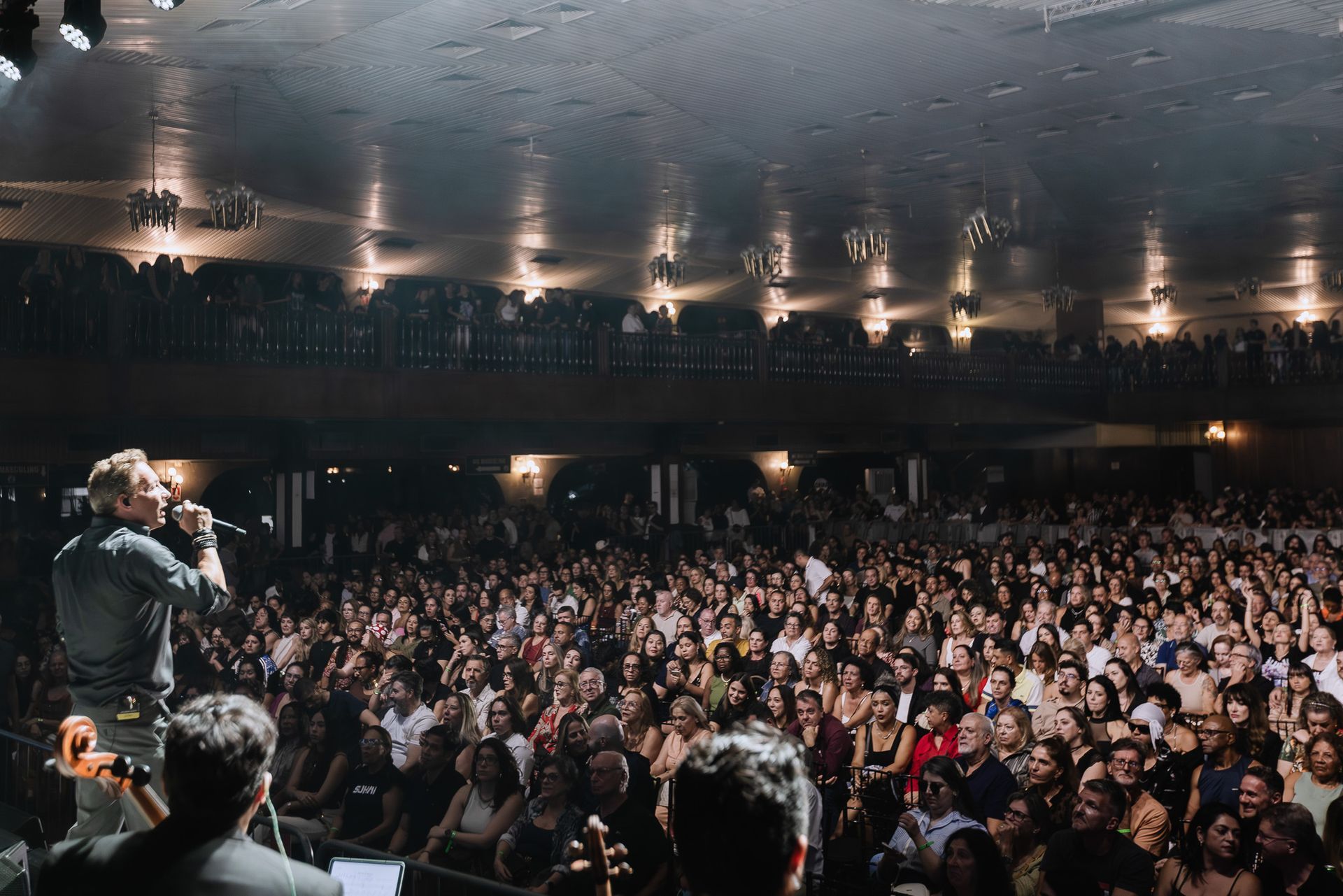 Músicos no palco de frente para uma grande plateia entusiasmada em uma sala de concertos com pouca luz.