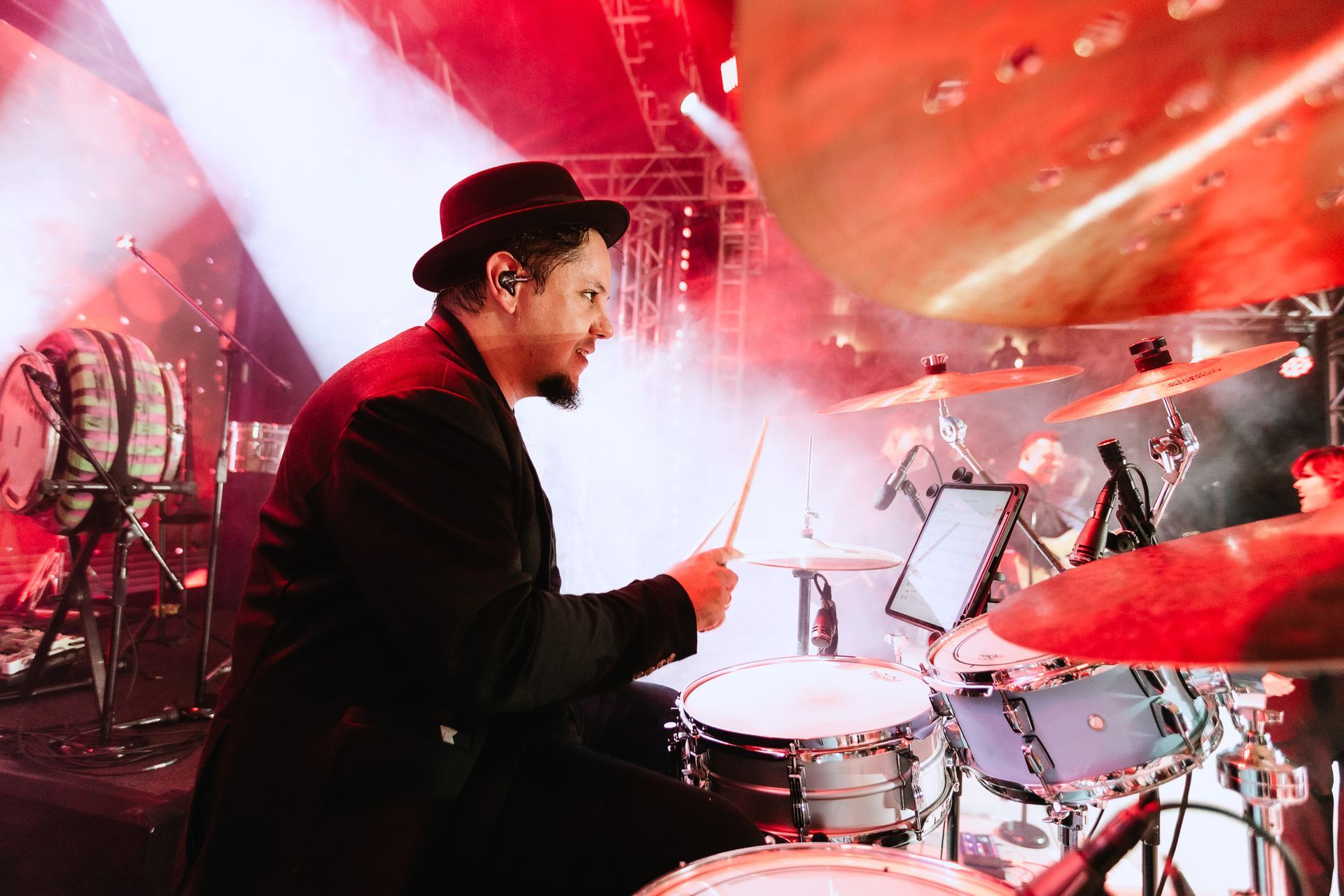 Baterista tocando em um palco iluminado em vermelho, com pratos e estantes de partituras por perto.