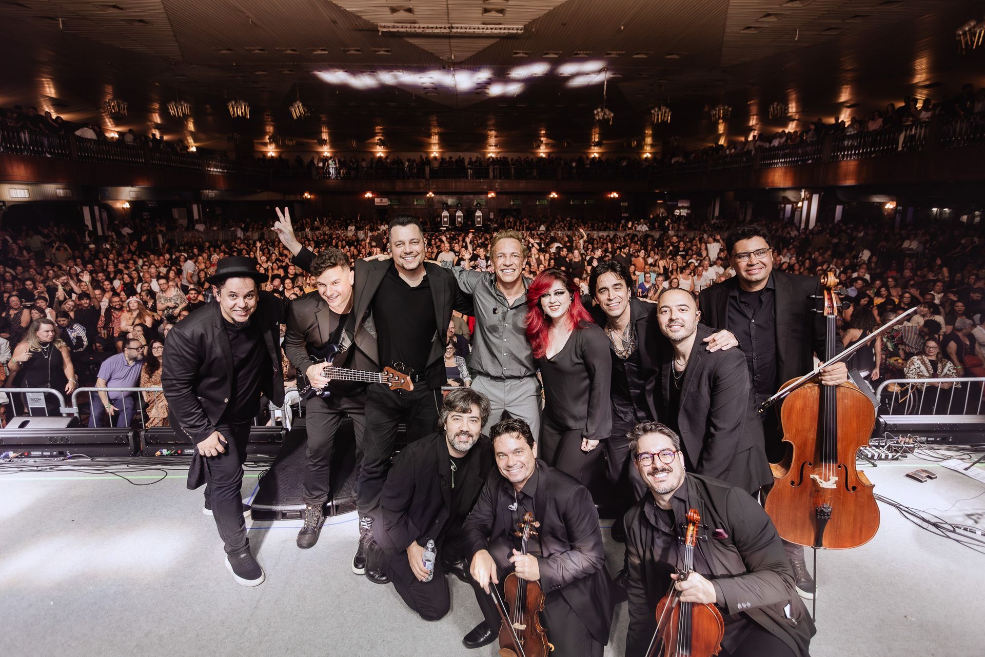 Membros da orquestra posando no palco de uma sala de concertos, com um violoncelo à direita e a plateia ao fundo.