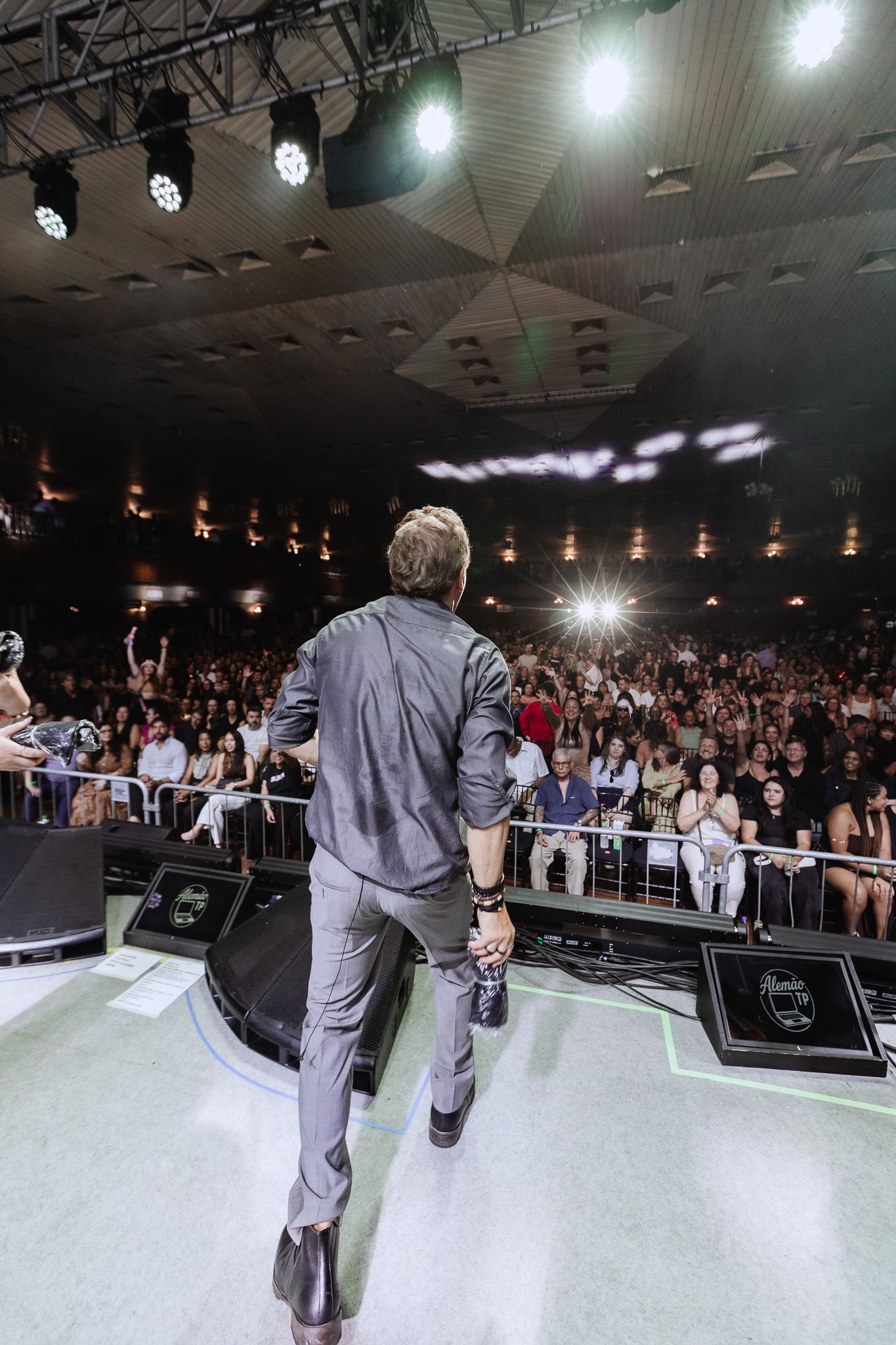 Artista no palco de frente para uma grande plateia em uma sala de concertos, com luzes brilhantes e alto-falantes no teto.