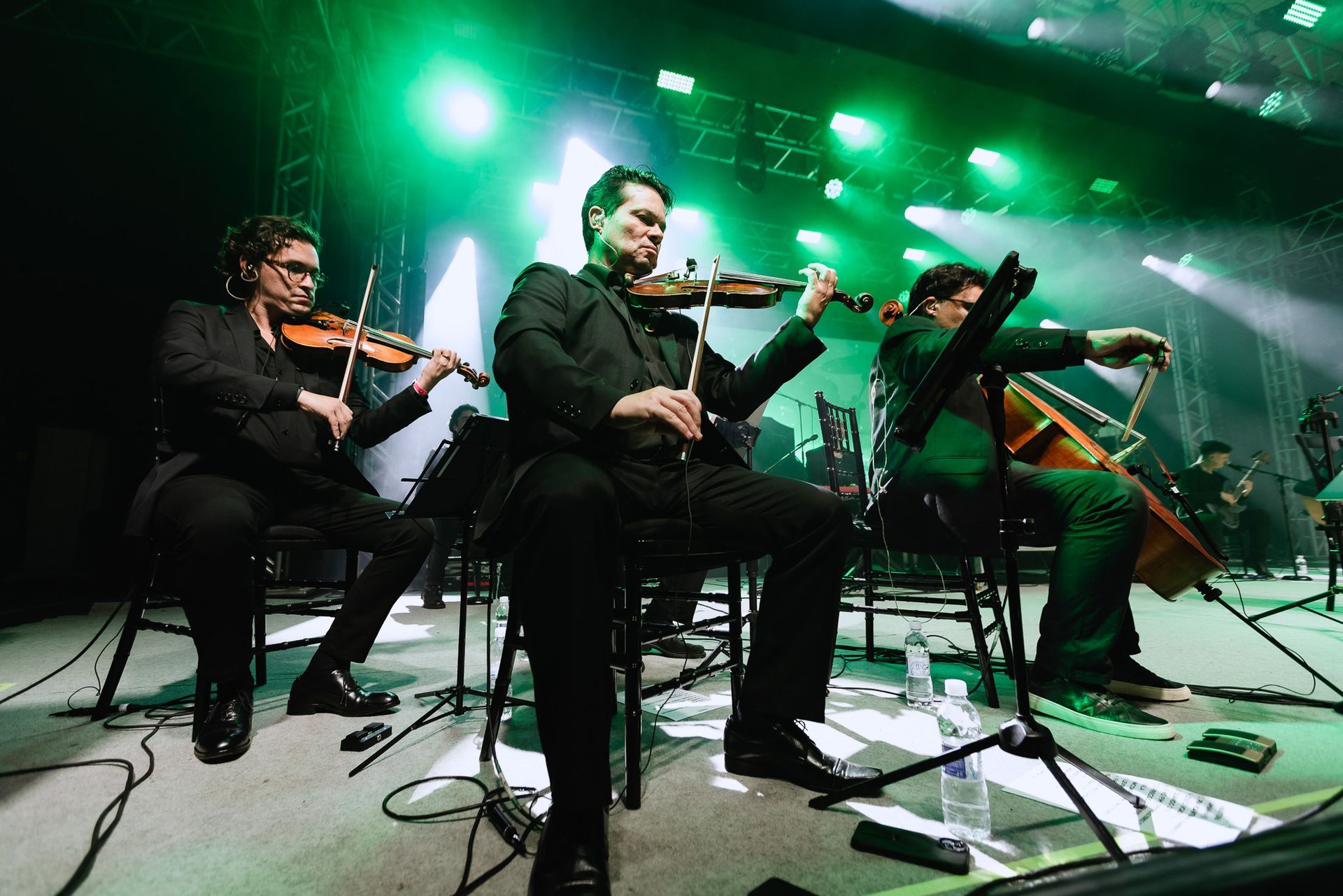 Três músicos tocando violinos no palco sob luzes verdes.