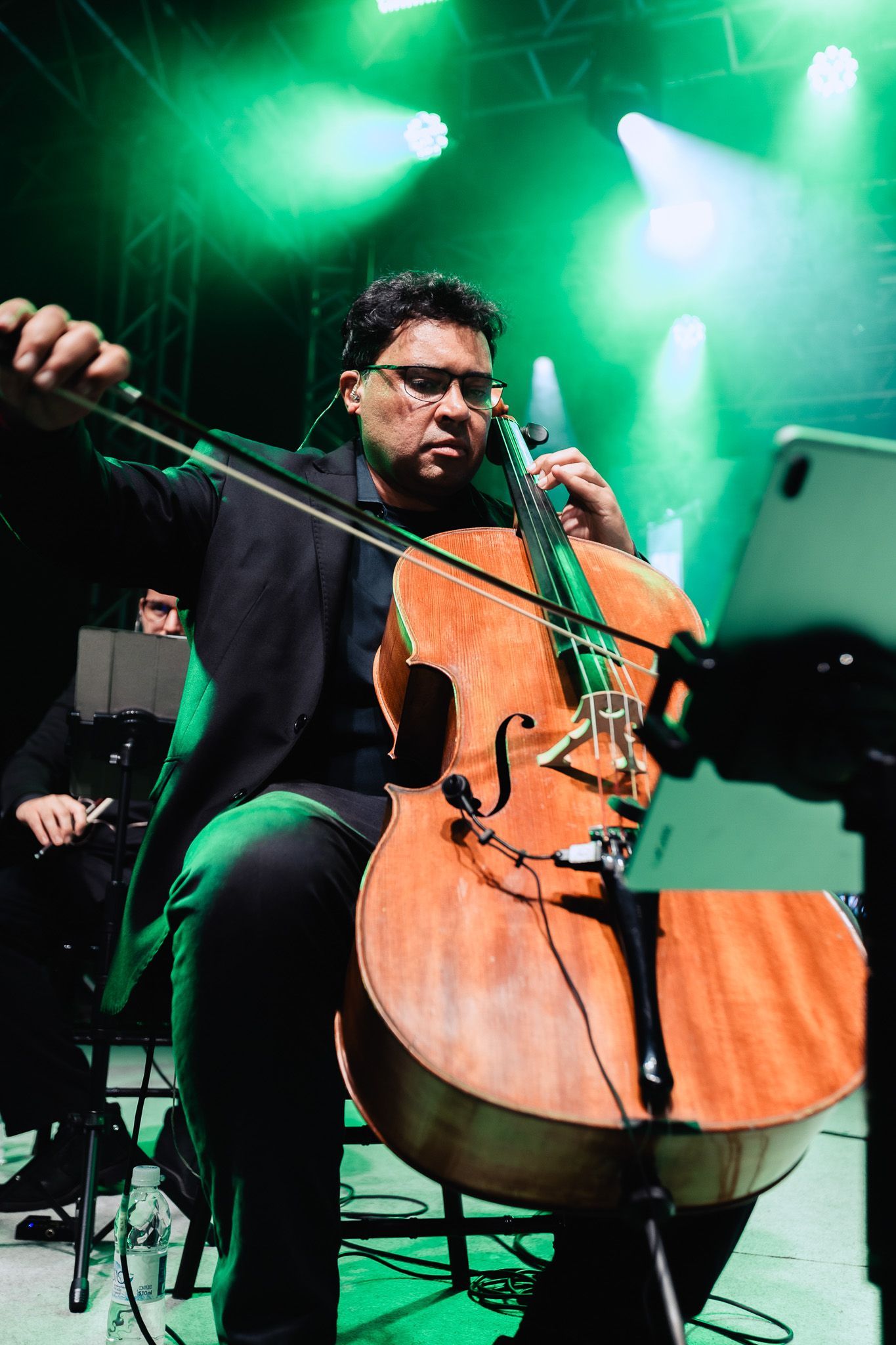 Violoncelista se apresentando no palco sob luzes verdes, sentada ao lado de uma estante de partituras.