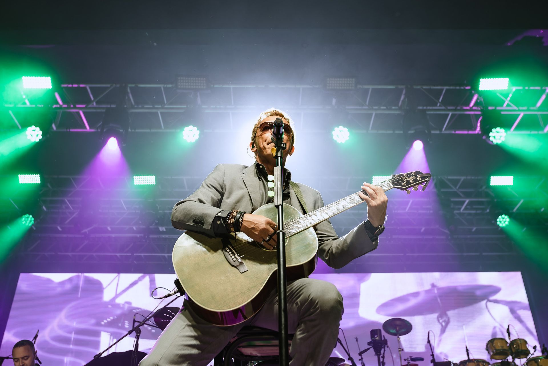 Cantor tocando violão em um palco iluminado com luzes roxas e verdes.