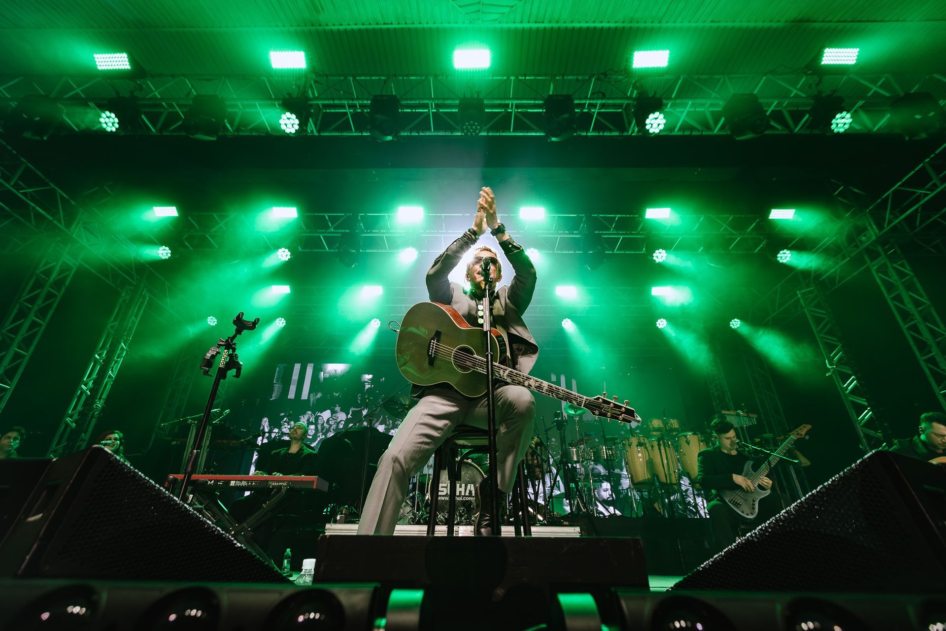 Banda de rock se apresentando no palco sob luzes verdes brilhantes, vocalista com os braços erguidos no centro do palco.