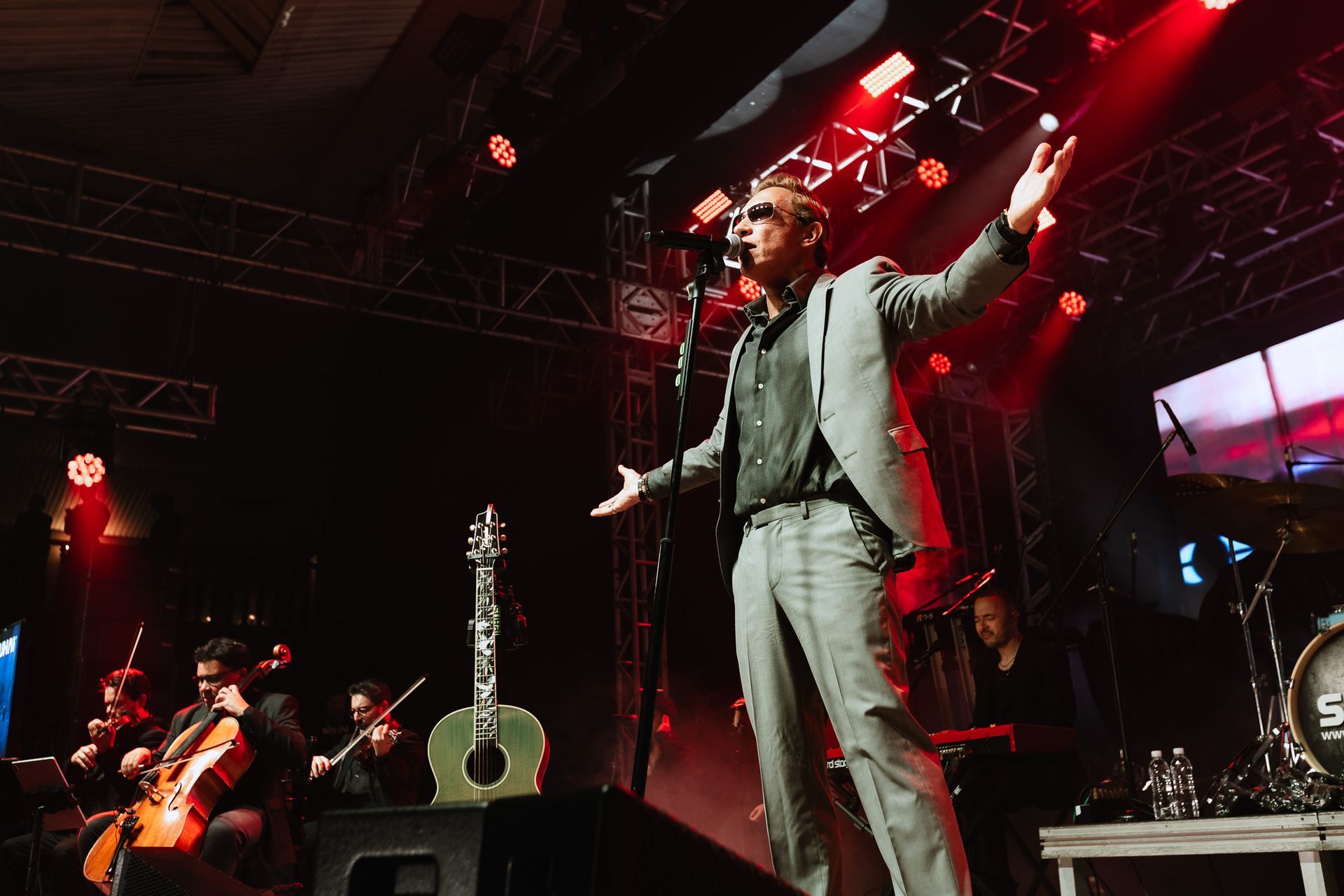 Cantor no palco com os braços estendidos sob luzes vermelhas, com um violão em um suporte próximo.