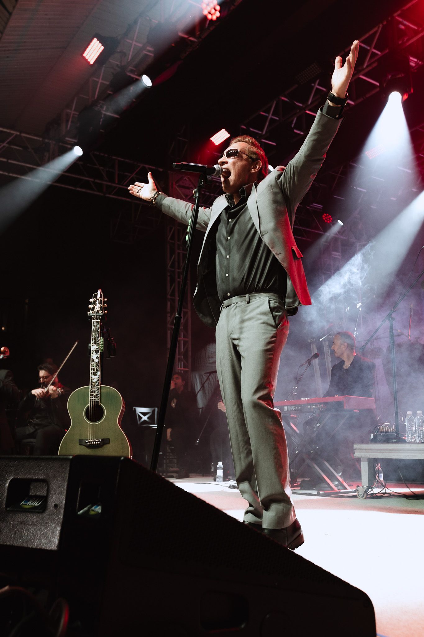 Cantor se apresentando no palco sob luzes vermelhas e brancas, com os braços erguidos ao lado de um violão.