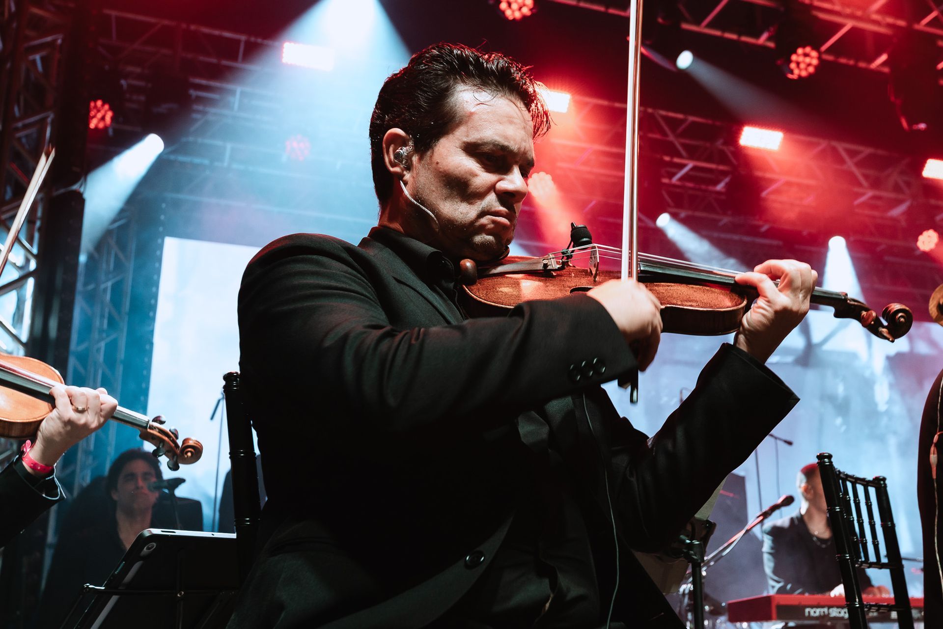 Violinista de terno preto se apresentando no palco sob luzes vermelhas e azuis.