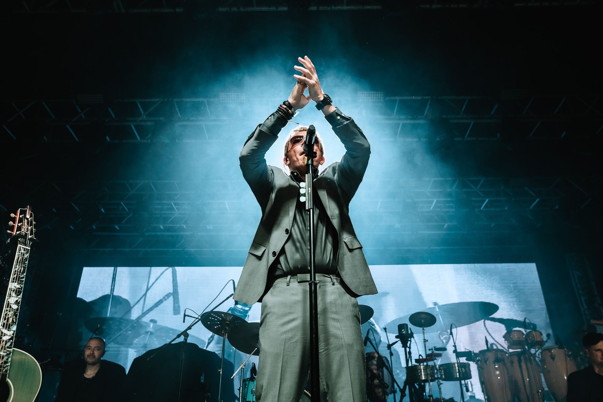 Singer onstage clapping under blue spotlight with band and drum kit in the background