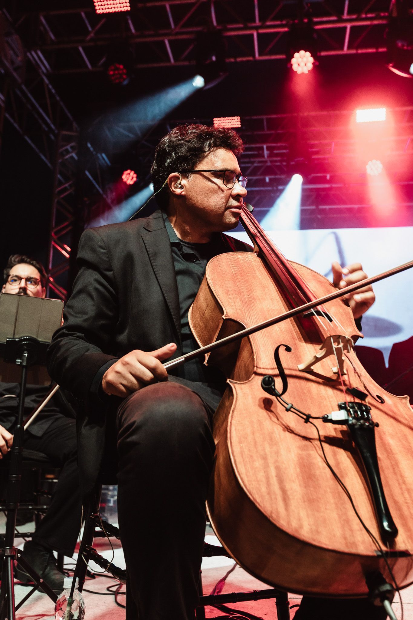 Violoncelista de terno preto se apresentando no palco sob luzes de concerto vermelhas e azuis.