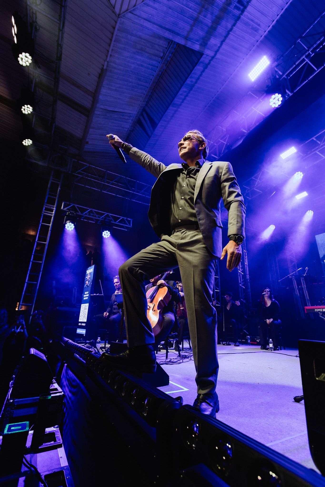 Cantor de terno escuro apontando para um palco de concerto iluminado em azul.