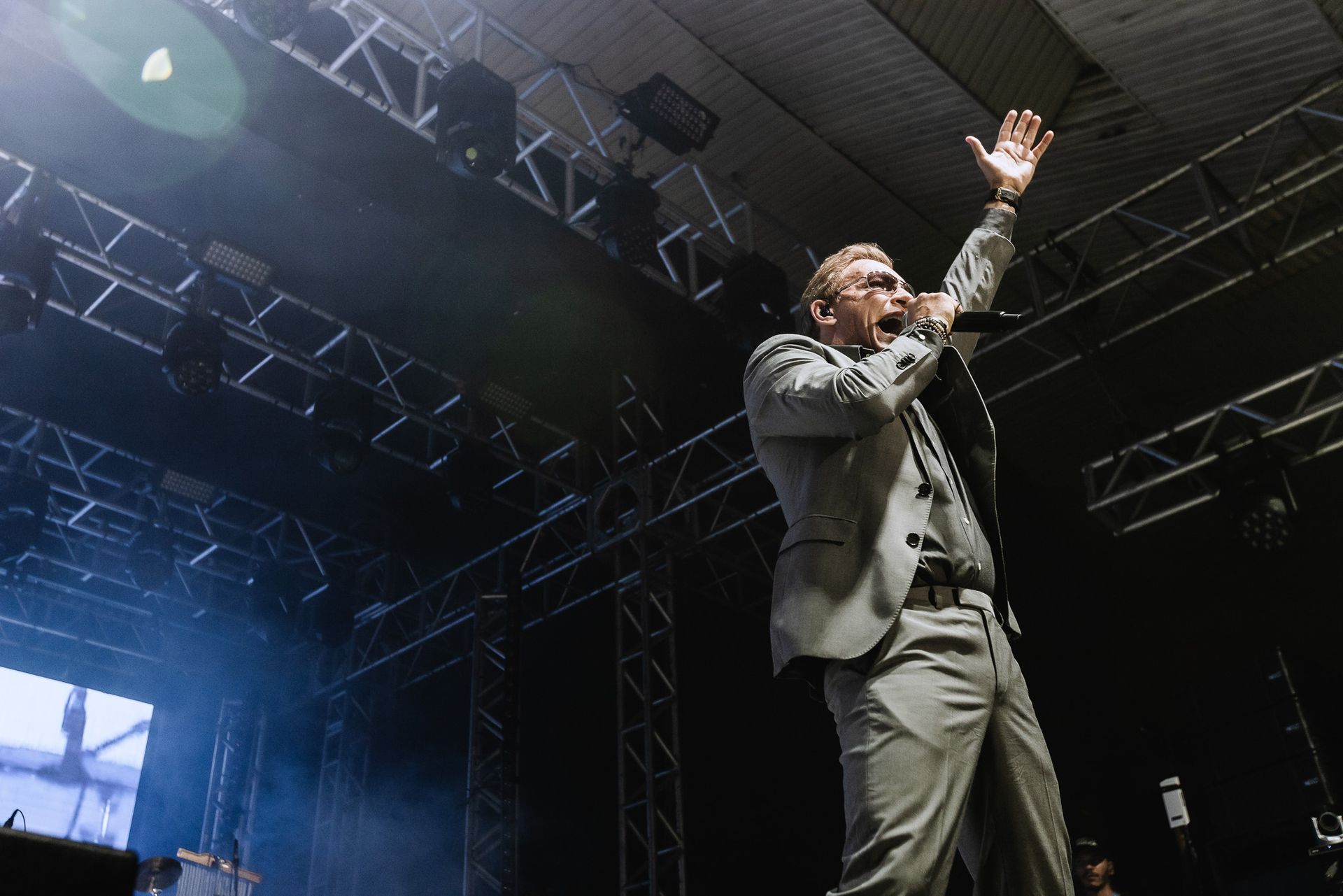 Singer in a gray suit performing onstage, raising one hand under bright concert lights.
