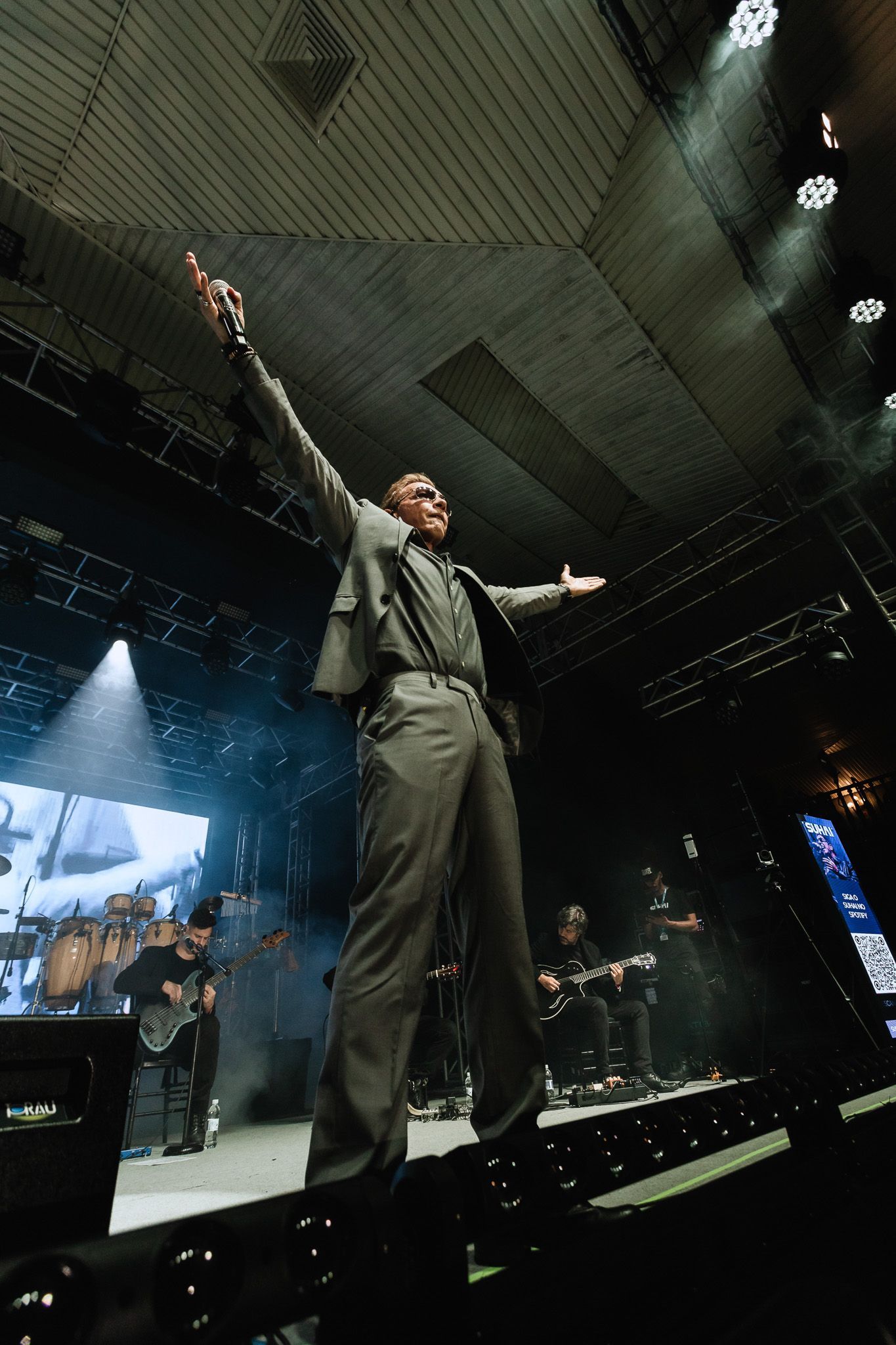 Cantor no palco com os braços erguidos sob luzes brilhantes de concerto; banda visível ao fundo.