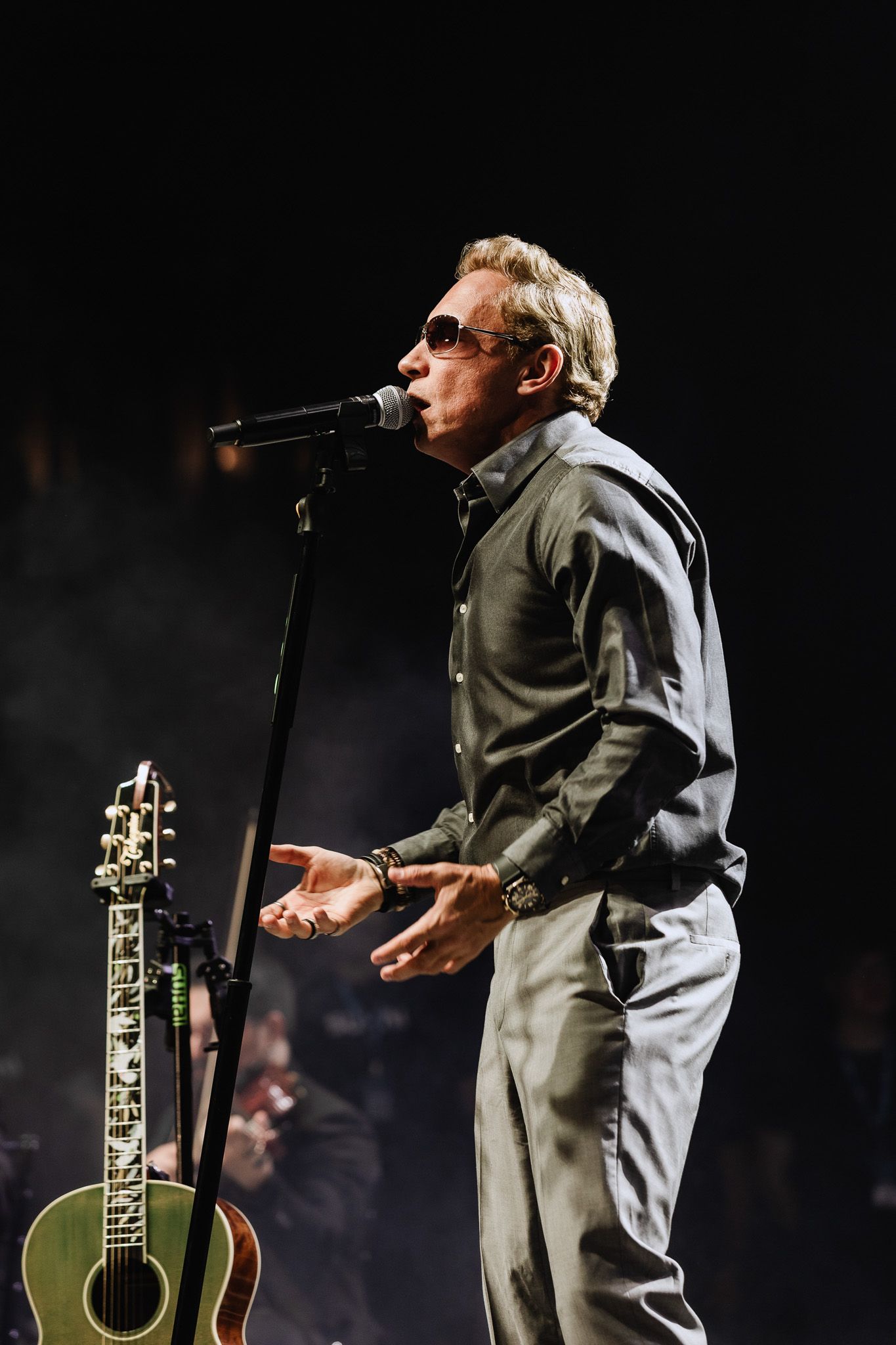 Cantor se apresentando no palco ao microfone, usando óculos escuros e uma camisa cinza, com um violão por perto.