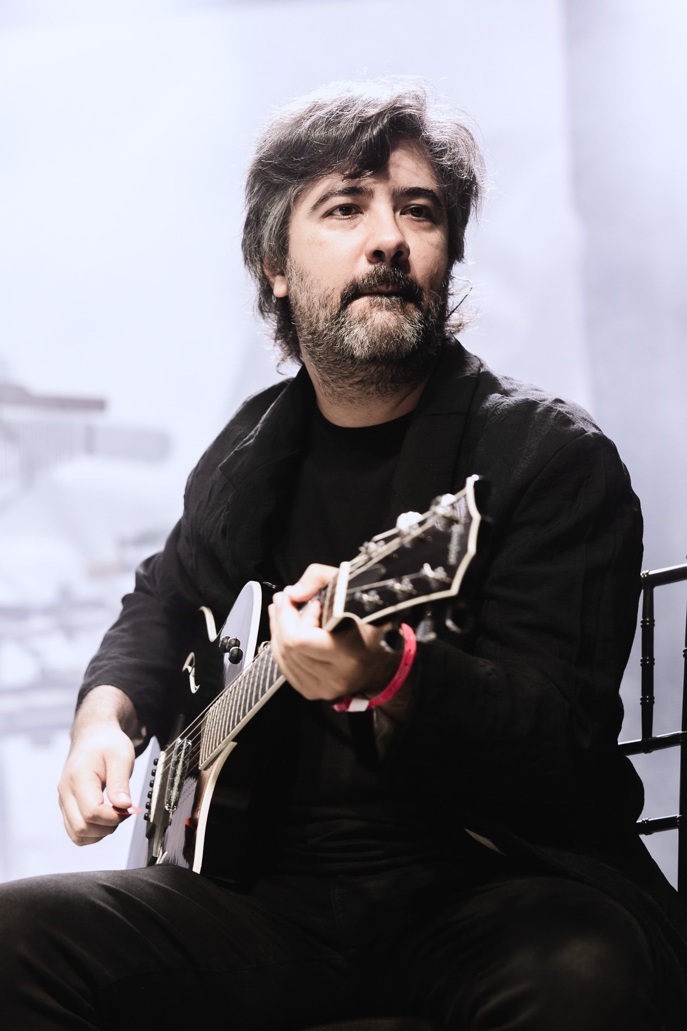 Músico sentado com uma guitarra elétrica, vestindo uma camisa preta contra um fundo de palco brilhante.