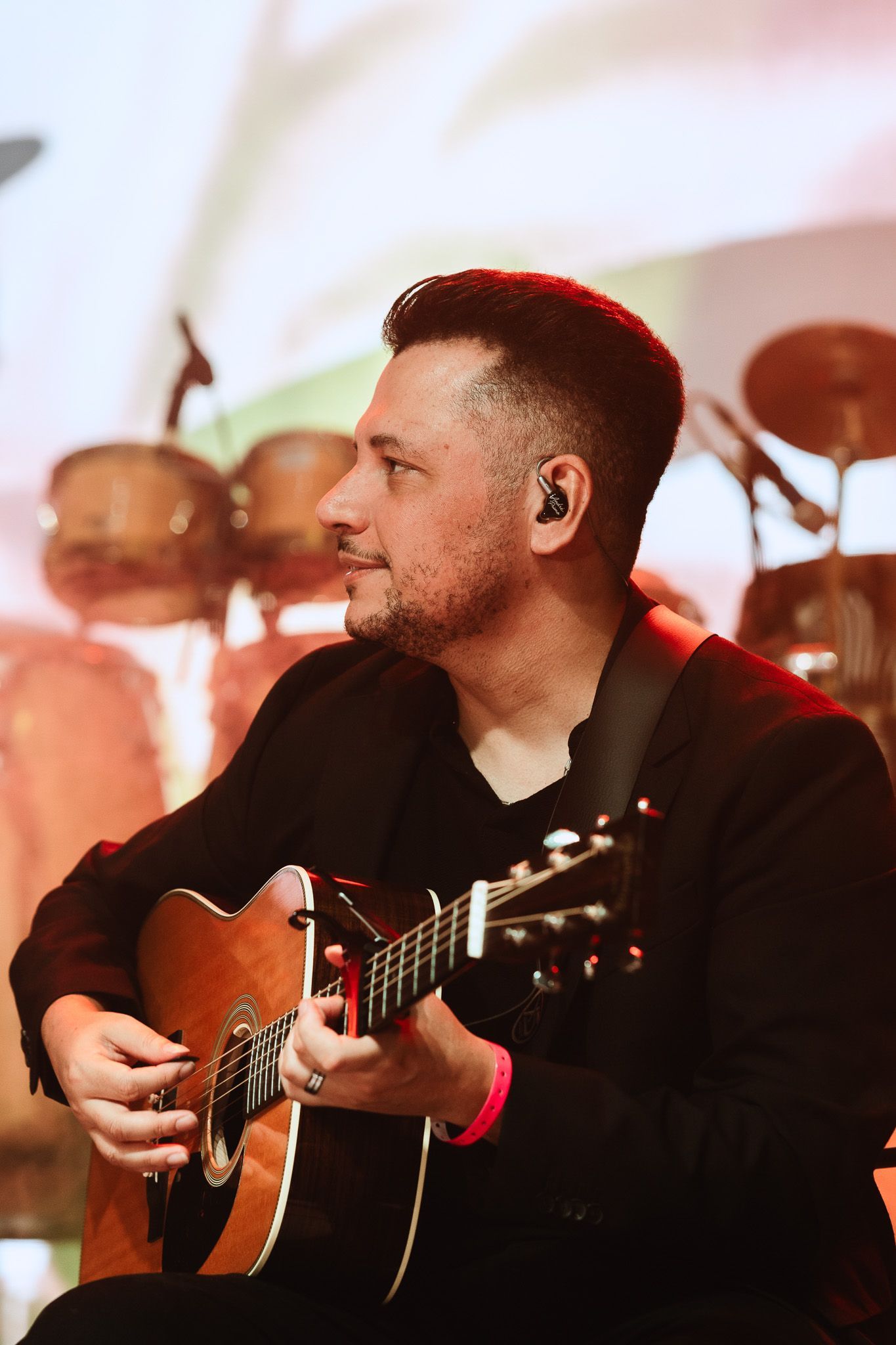 Músico tocando violão no palco, vestindo uma camisa preta, com bateria desfocada ao fundo.