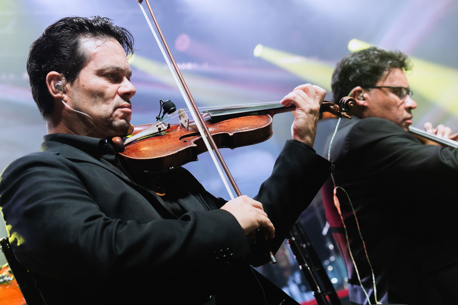 Dois violinistas se apresentando no palco sob luzes coloridas de concerto.