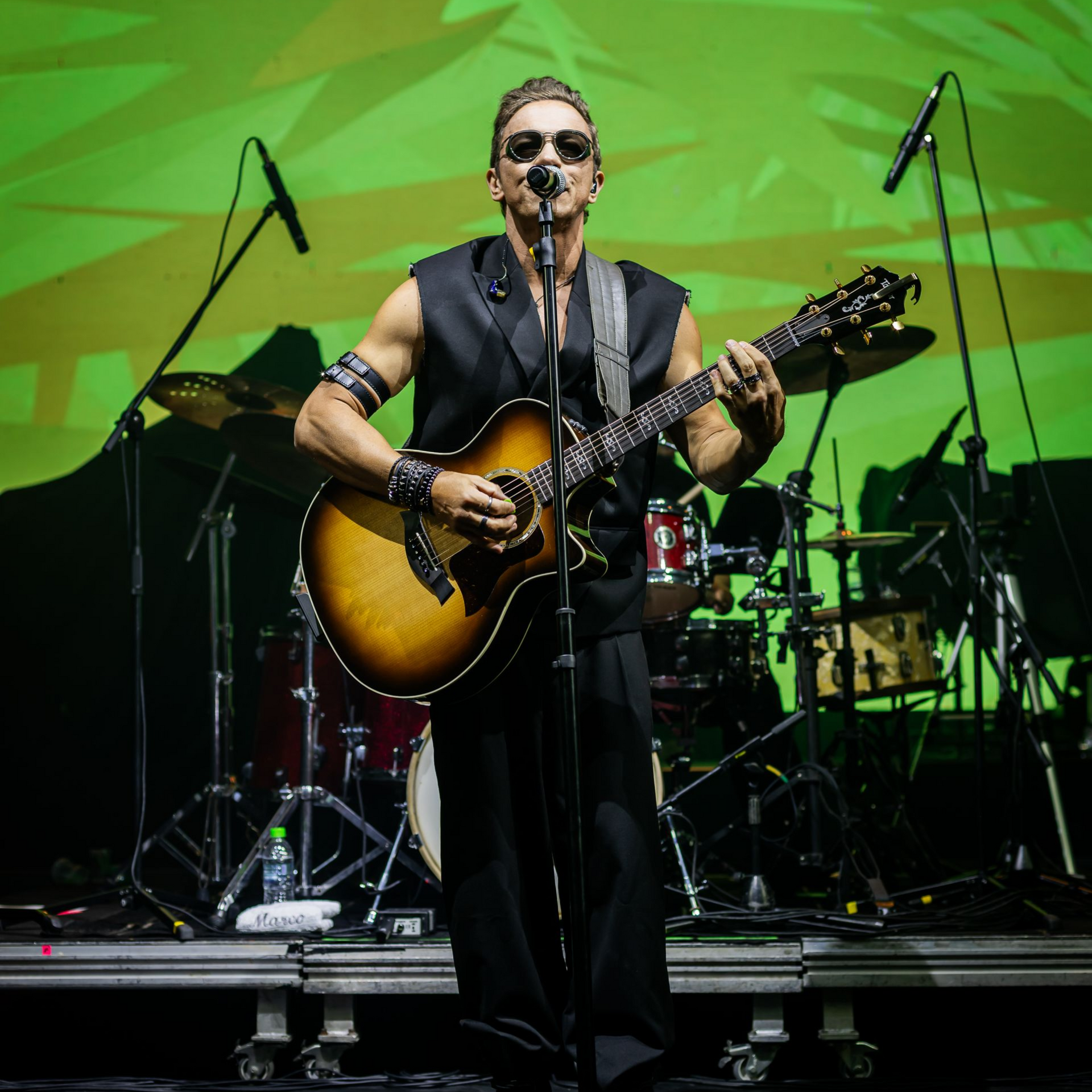 Cantor tocando violão no palco com luzes verdes e uma bateria ao fundo.