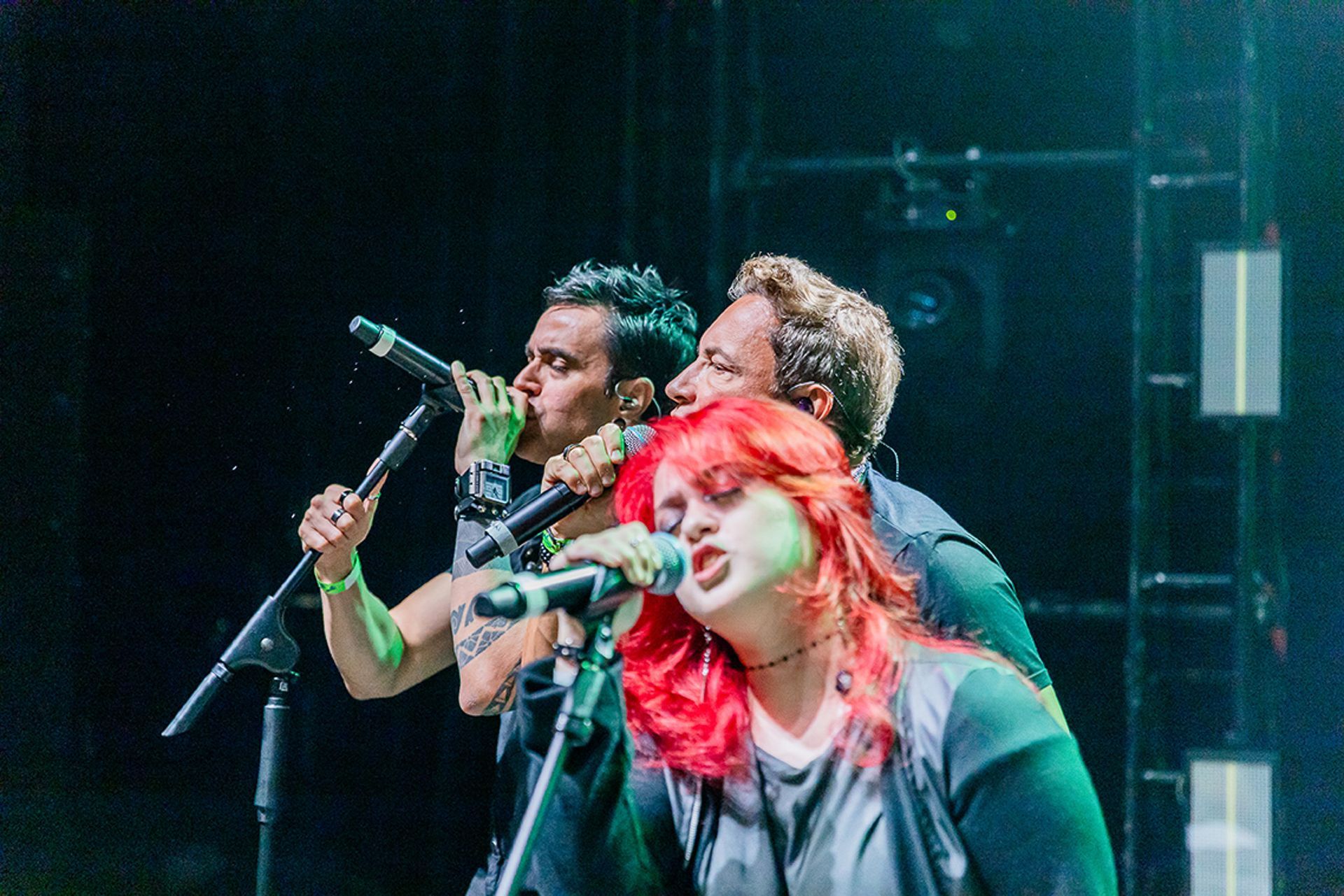 Três cantores se apresentando no palco, com uma cantora ruiva à frente, ao microfone.