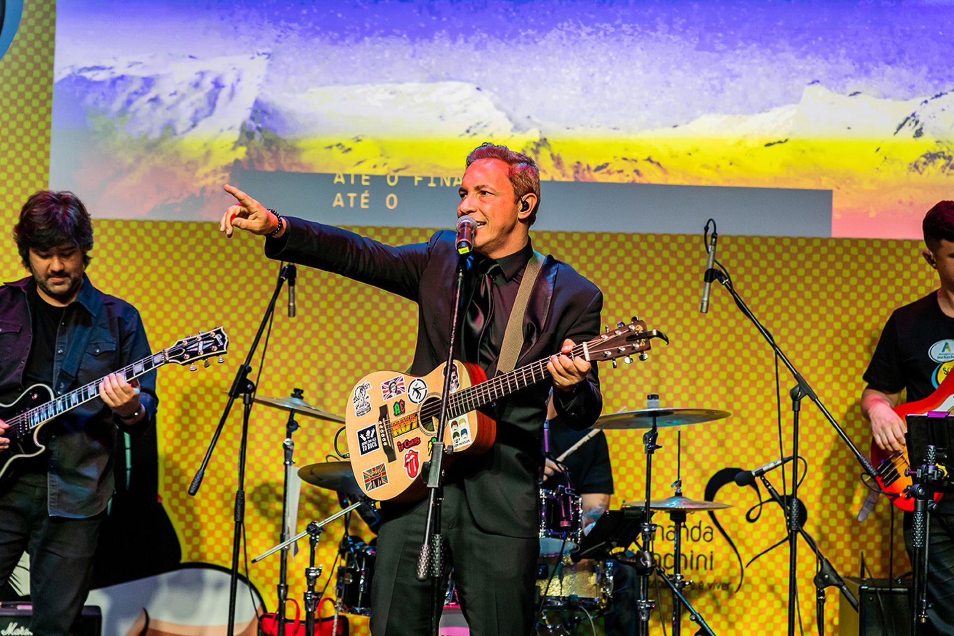 Um artista de terno preto toca um violão coberto de adesivos e aponta enquanto se apresenta no palco com uma banda.