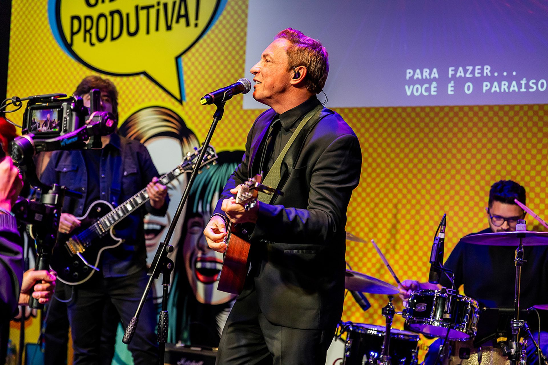 Um cantor tocando violão no palco com um guitarrista e um baterista, tendo um fundo amarelo com texto em estilo de quadrinhos