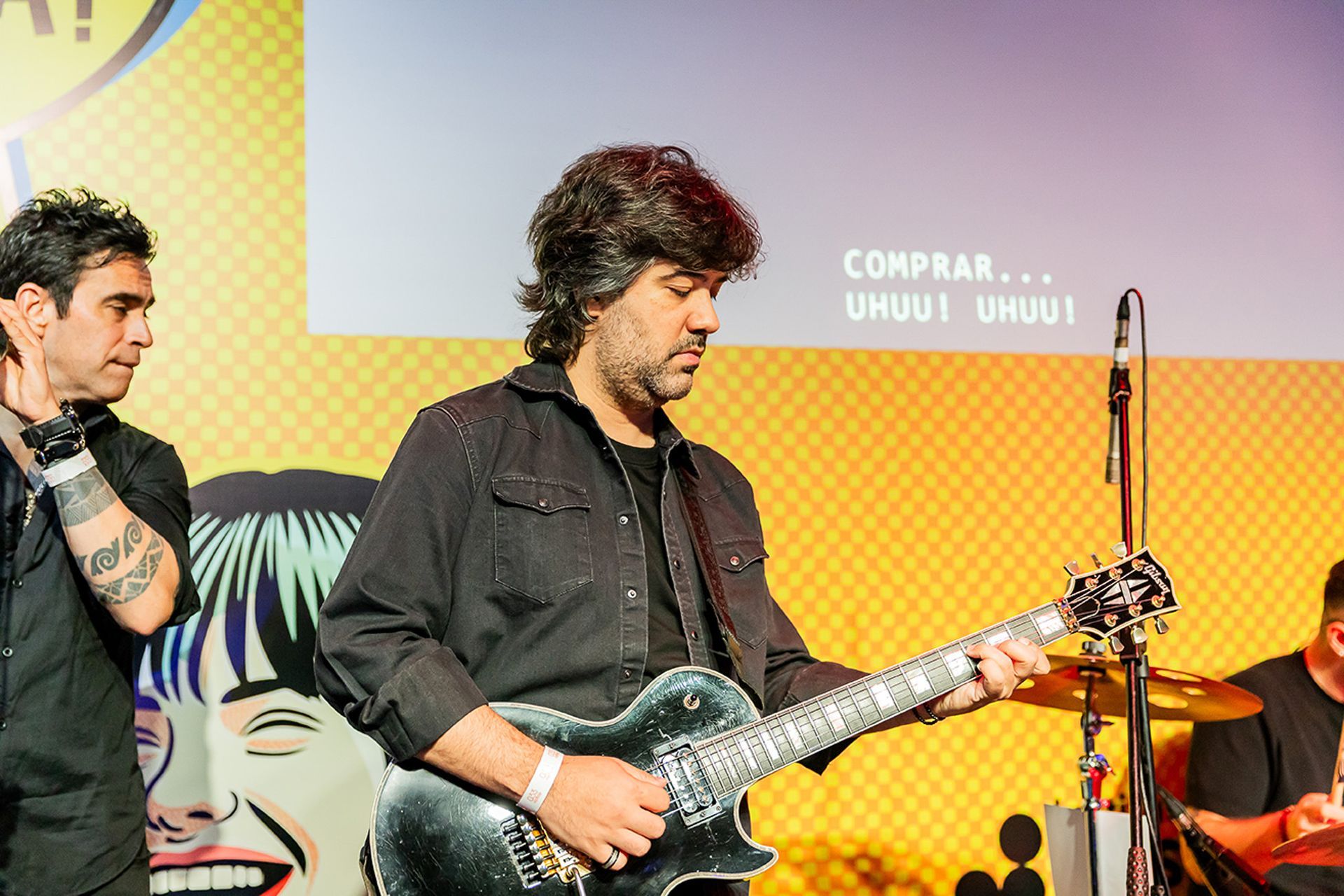 Um músico toca uma guitarra elétrica escura no palco, em frente a um cenário colorido e estampado.