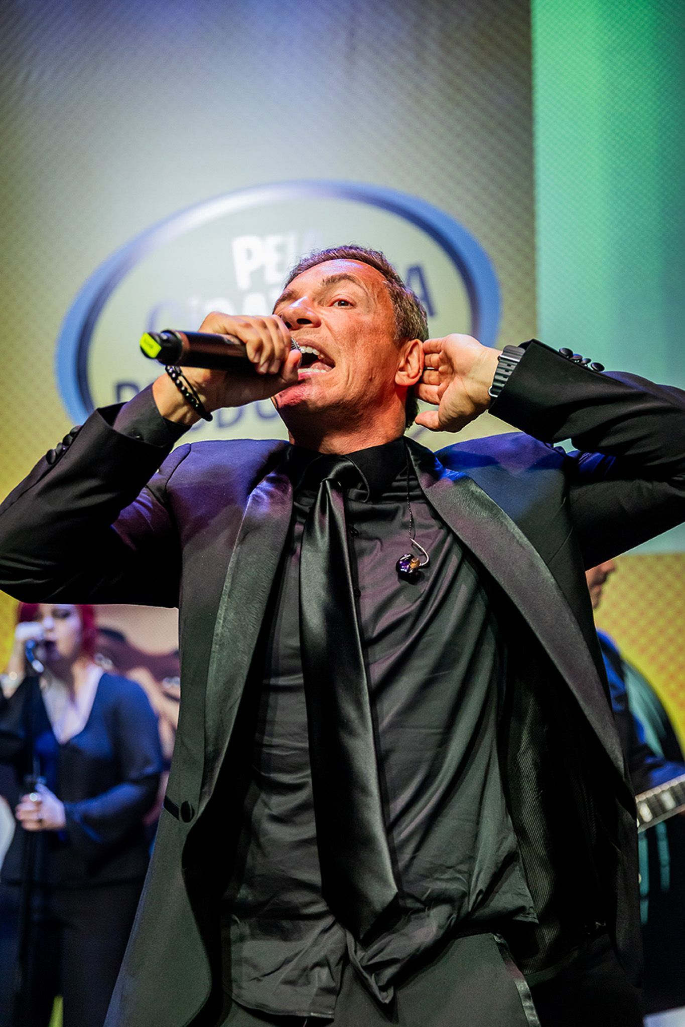 Um cantor de terno e gravata pretos canta intensamente em um microfone no palco, com um músico visível ao fundo.