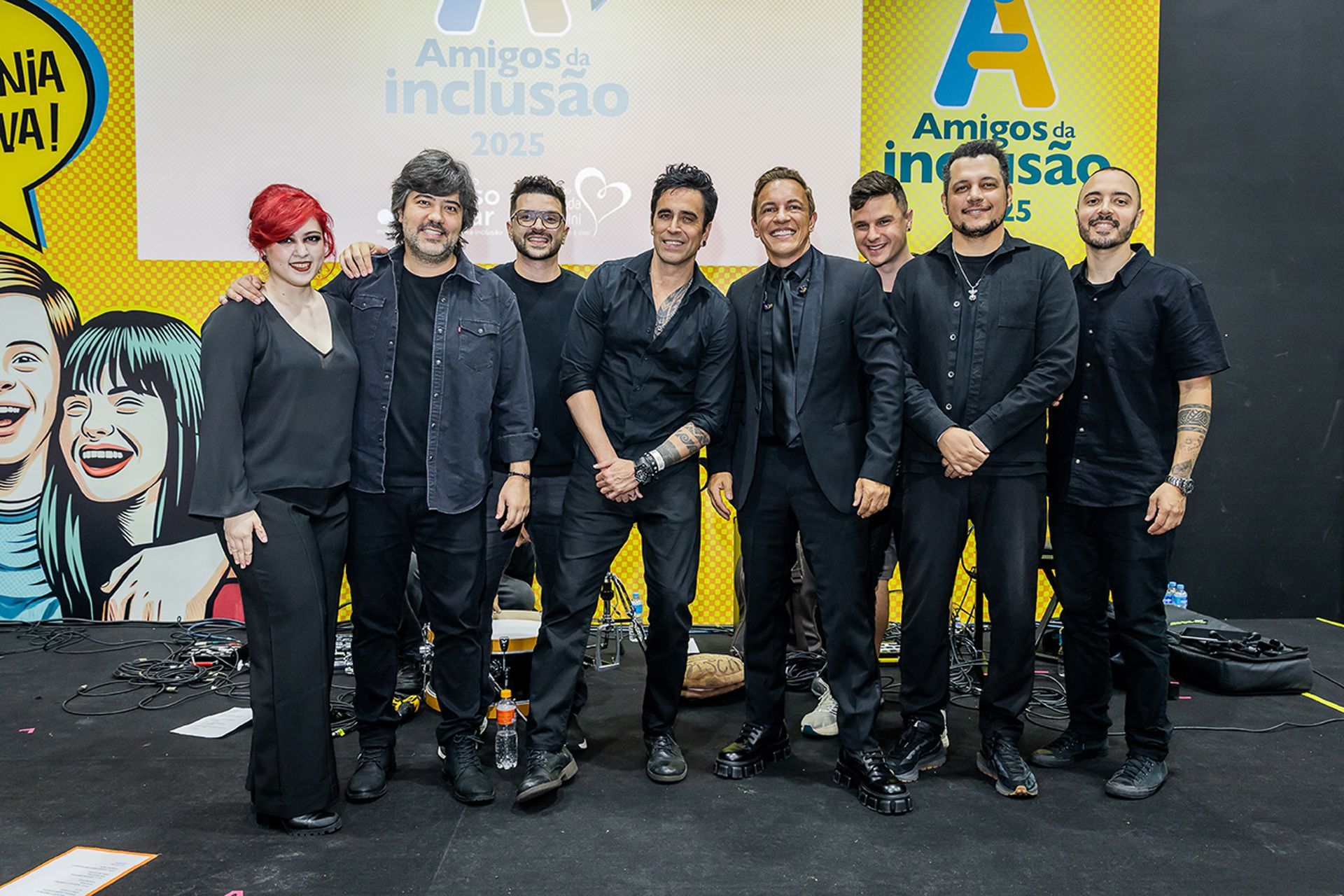 Sete pessoas vestidas de preto posando em um palco com um painel de fundo amarelo brilhante escrito “Amigos Inclusão”.