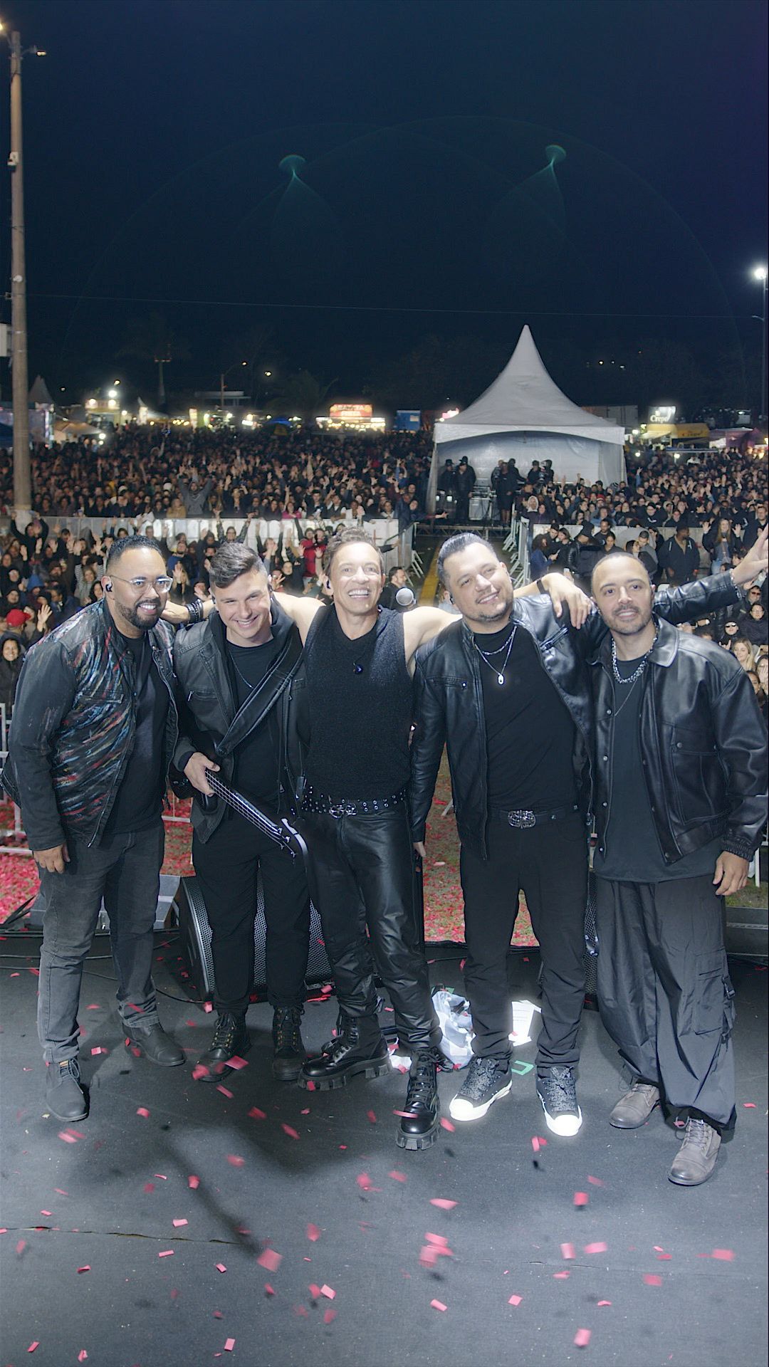 Seis pessoas vestidas de preto em um palco de shows à noite, sorrindo, com uma grande multidão e uma tenda ao fundo.