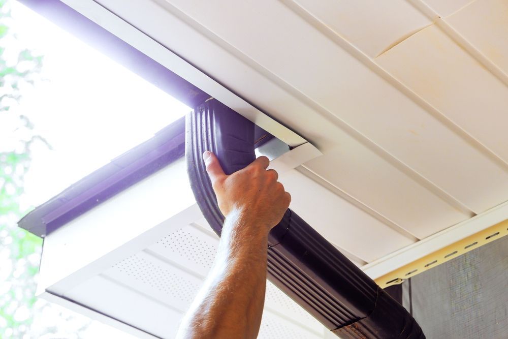Hand installing a black gutter section on a white house under an eave.
