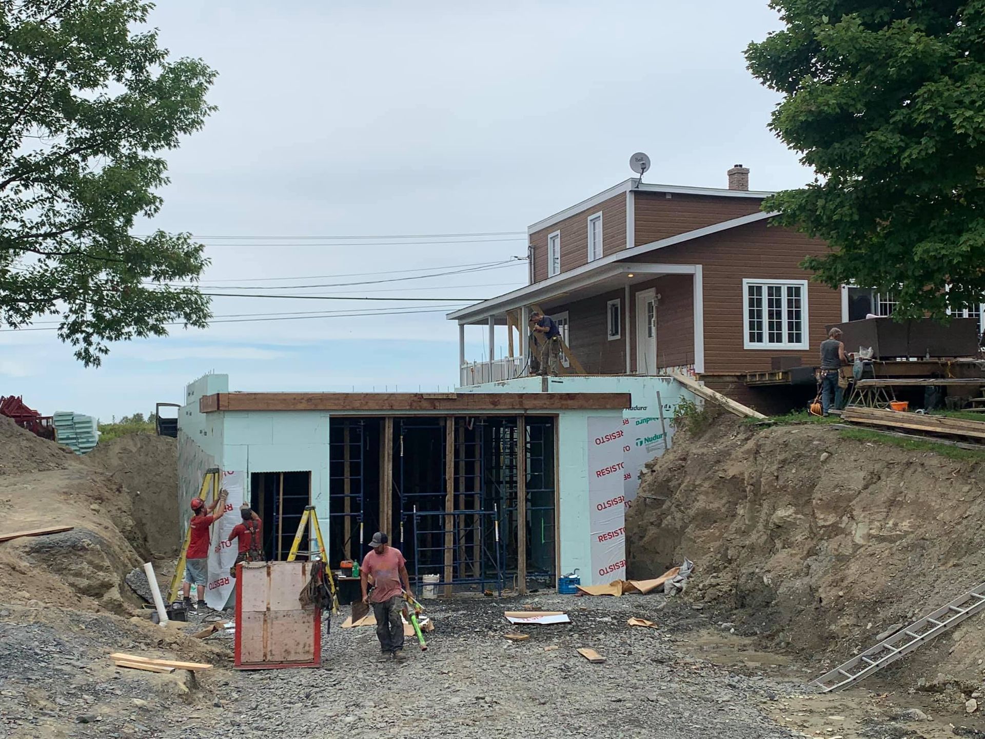 Chantier : ouvriers construisant une structure adjacente à une maison brune à deux étages. Zone excavée, ciel nuageux.