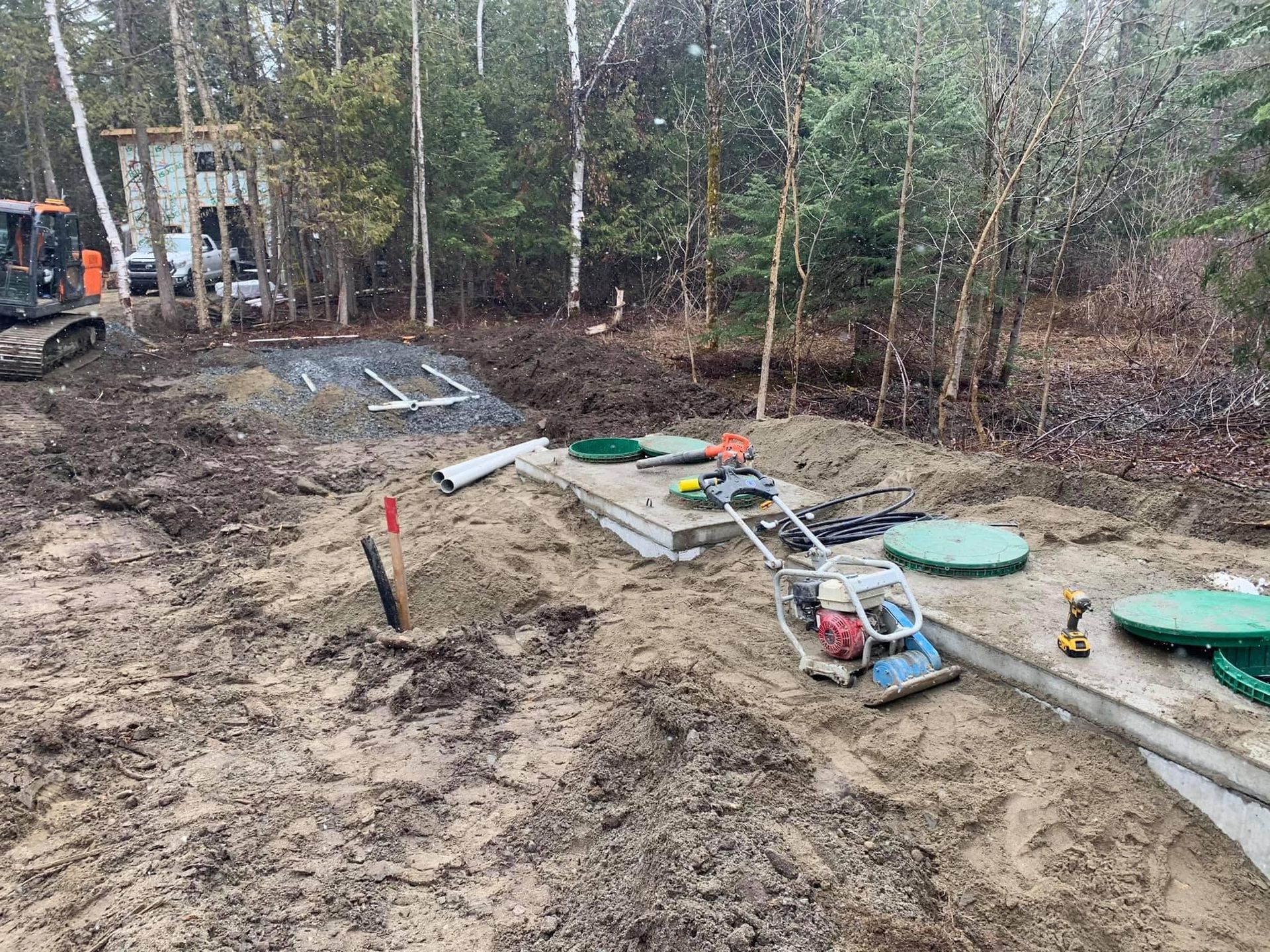 Chantier de construction avec fosses septiques, compacteur et mini excavatrice dans un secteur boisé.