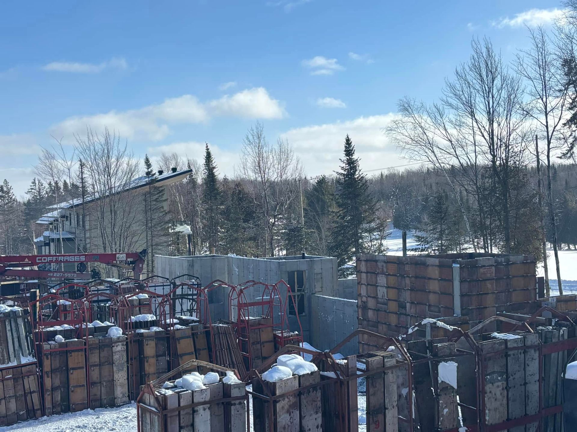 Scène d'hiver : Bâtiment et matériaux de construction entourés de neige, avec des arbres et un ciel bleu vif.