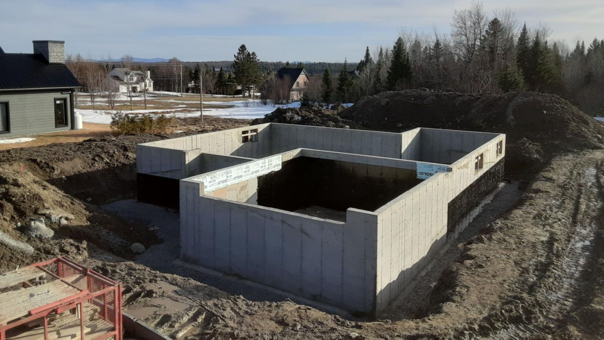 Fondation en béton d'une maison en construction, entourée de terre, avec des maisons et des arbres en arrière-plan.