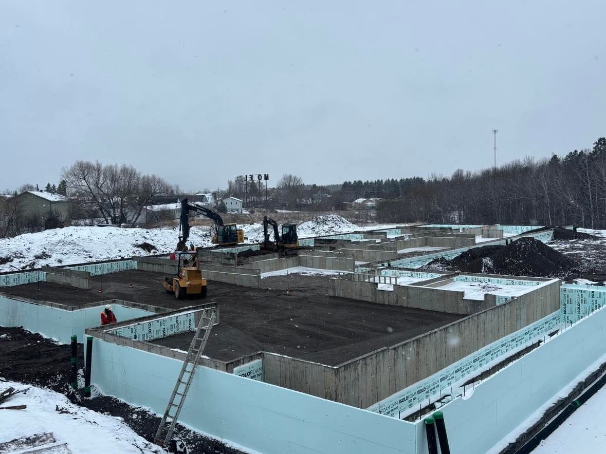 Chantier en hiver : murs de fondation, engins, sol enneigé, ciel couvert.