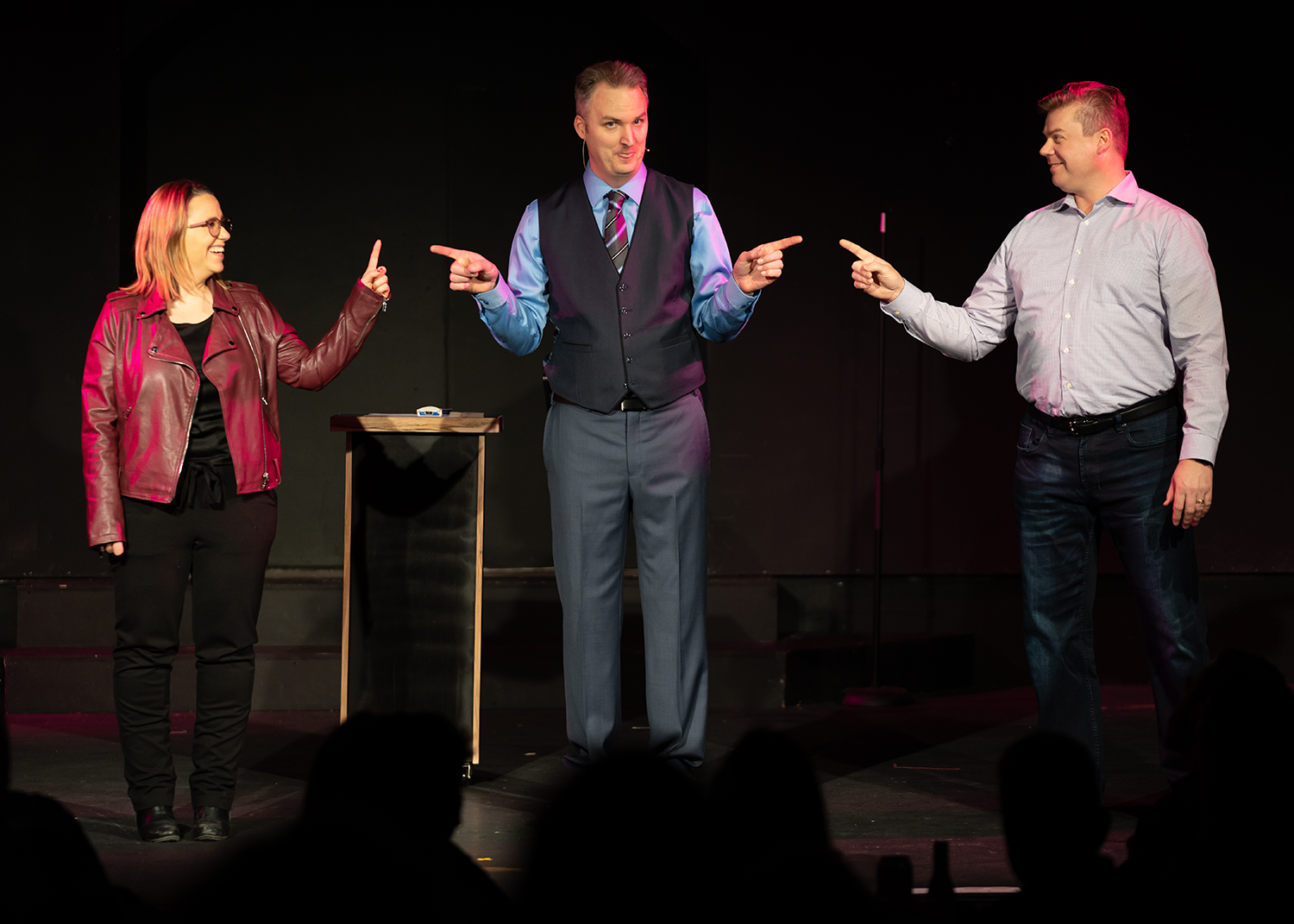 Three people are standing on a stage pointing at each other.
