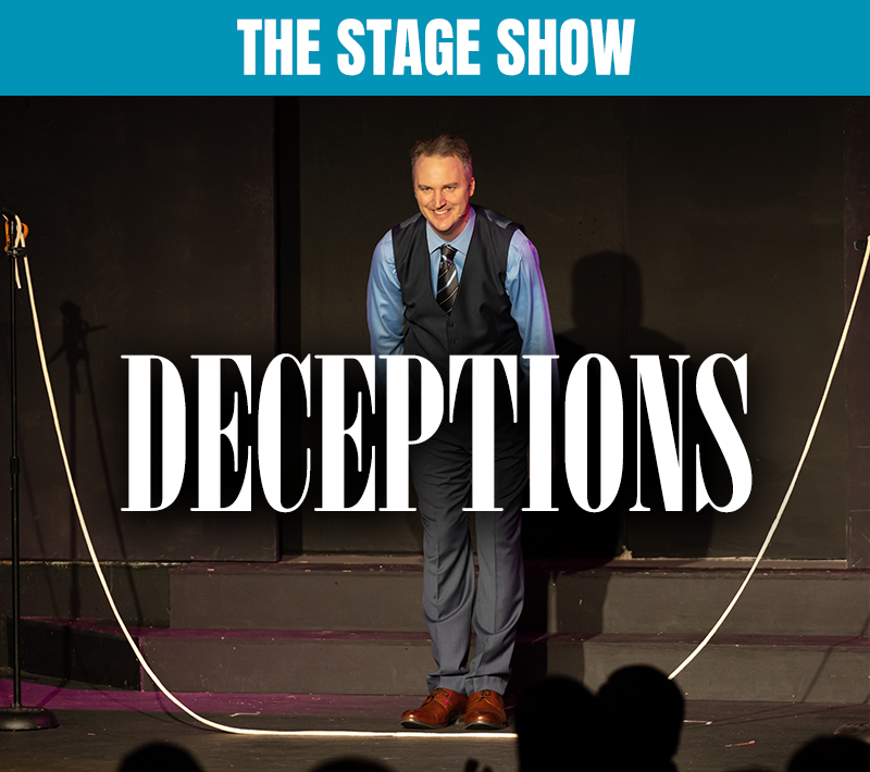 A man in a suit and tie stands on a stage in front of a crowd and a sign that says the stage show deceptions
