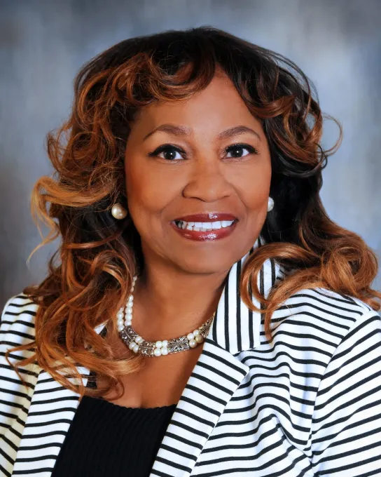 A smiling professional wearing a striped blazer and a pearl necklace against a mottled gray background.