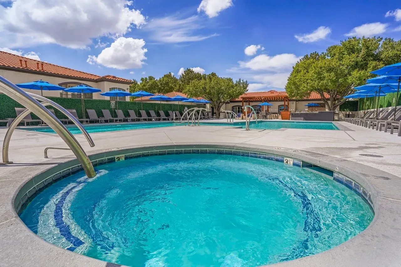 Outdoor swimming pool and jacuzzi area with lounge chairs and blue umbrellas at La Ventana, offering apartments in Albuquerque, NM.