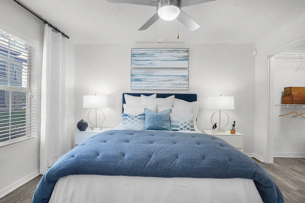 A bright and airy bedroom featuring a large blue comforter, a navy headboard, two white nightstands with lamps, and a window with blinds at La Ventana, offering apartments in Albuquerque, NM.