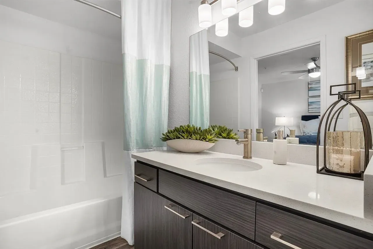 Modern bathroom with a white countertop, sink, and brushed nickel faucet at La Ventana, offering apartments in Albuquerque, NM. A shower/tub combo with a light blue and white curtain is visible .