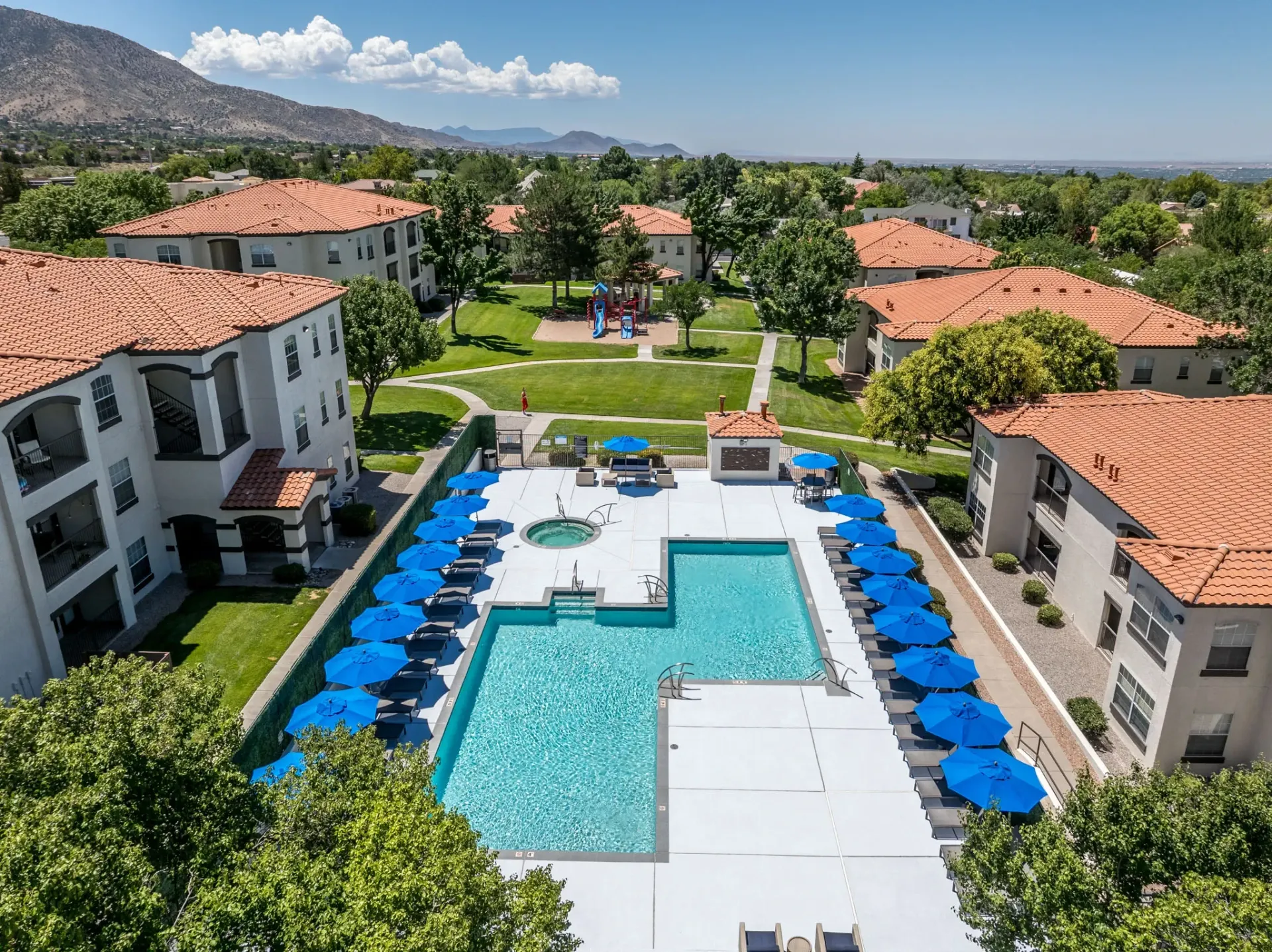 Aerial view of a resort-style swimming pool and spa area with lounge chairs and blue umbrellas at La Ventana, offering apartments in Albuquerque, NM.