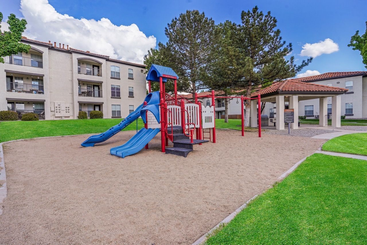 Playground with blue slides and red climbing structure in a grassy apartment community courtyard.