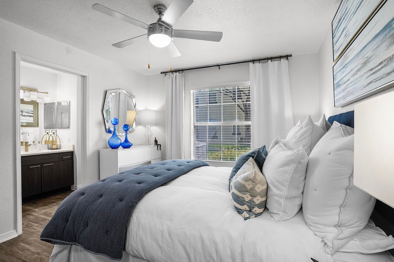 Bright bedroom with white walls, large window, ceiling fan, white bedding, and blue tufted quilt.