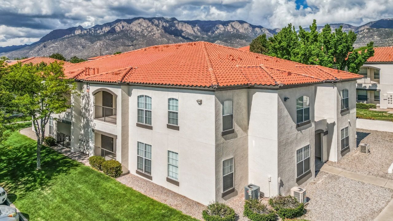 Exterior view of a beige apartment building with a red-tile roof, green lawn, and mountain backdrop.