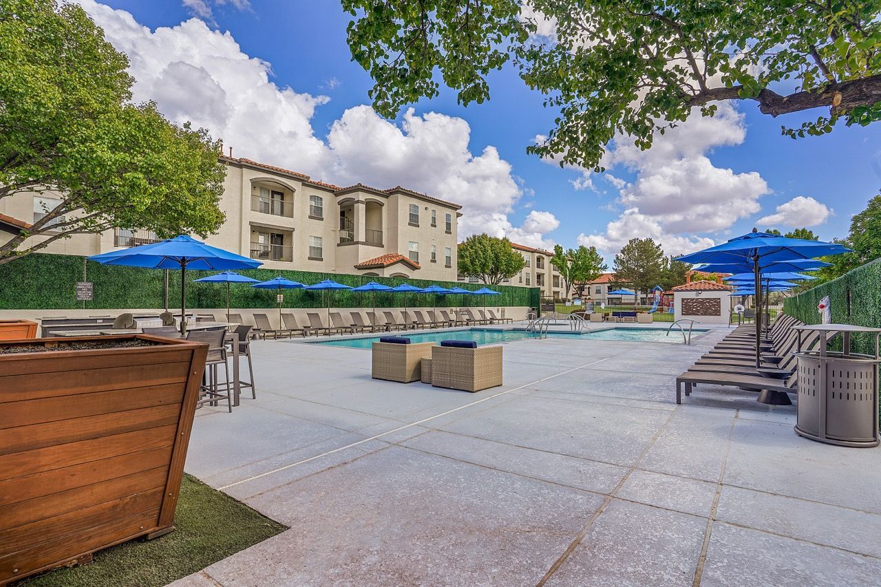 Outdoor community pool area with lounge chairs, blue umbrellas, and surrounding apartment buildings.