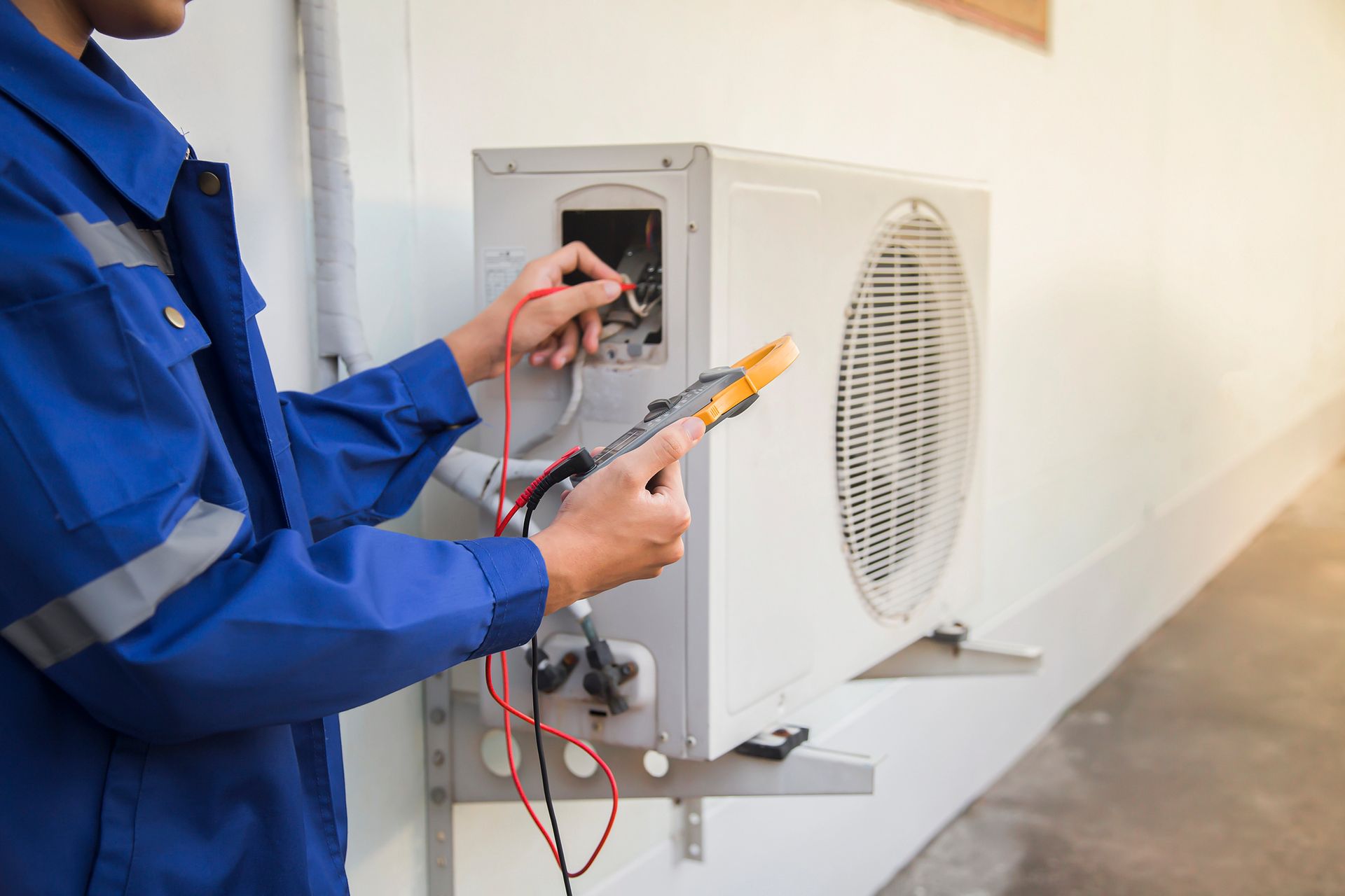 Un tecnico HVAC in tuta blu testa un'unità di aria condizionata con un multimetro.