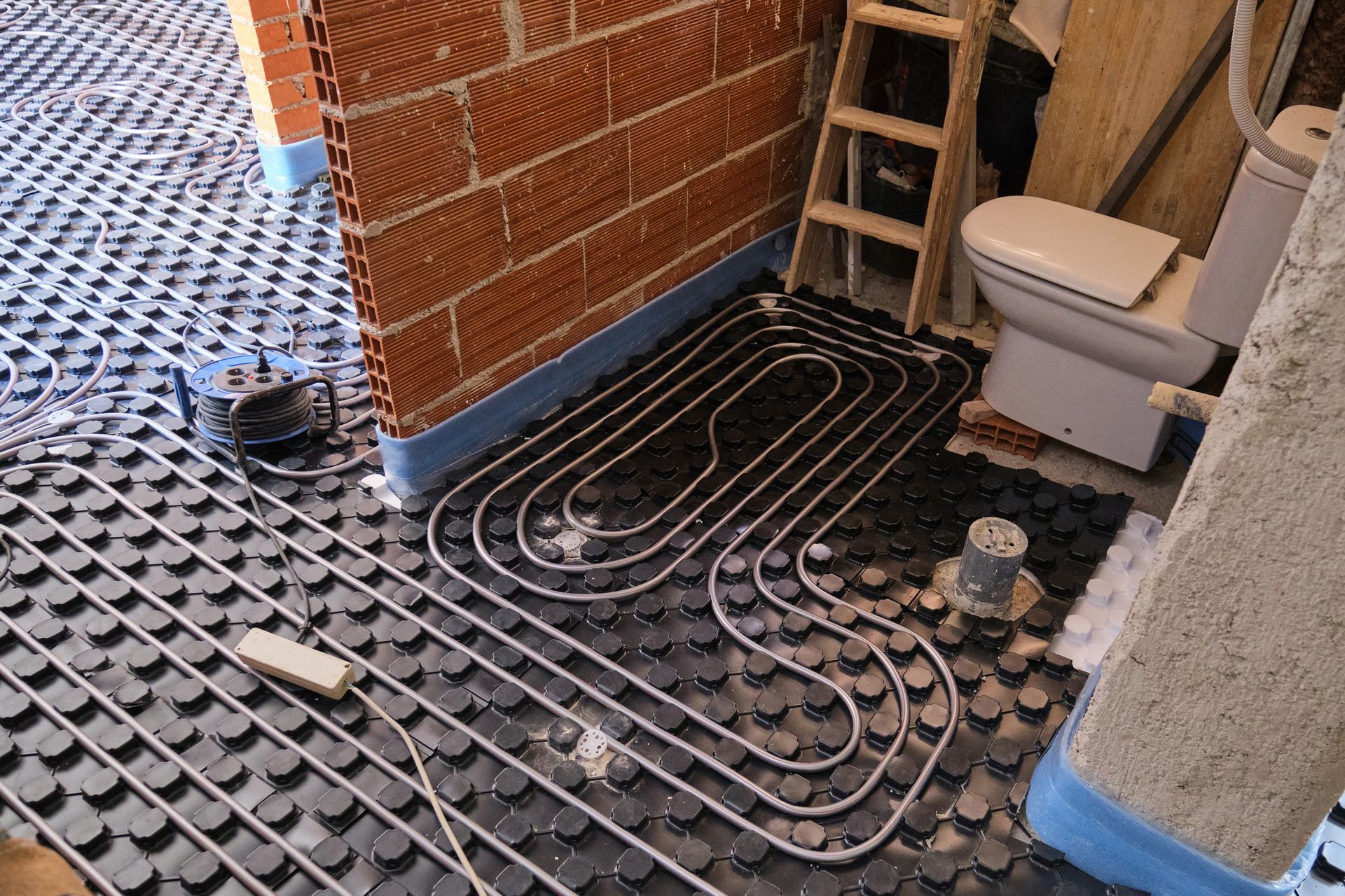 Installazione del riscaldamento a pavimento: tubi a spirale su isolamento nero, accanto a un muro di mattoni e al water.